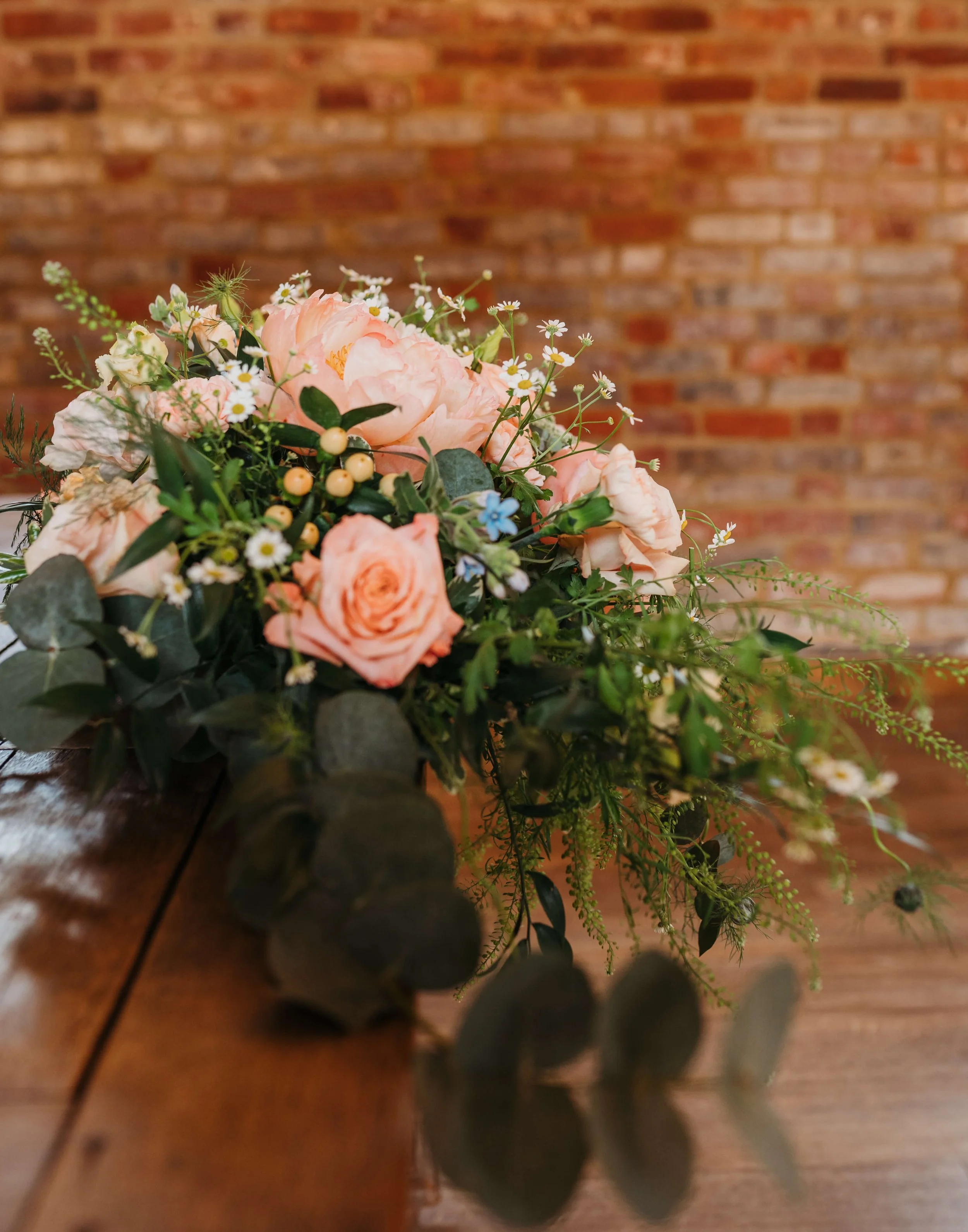 A bouquet of pink roses, white daisies, small blue flowers, and green foliage resting on a wooden table with a brick wall background.
