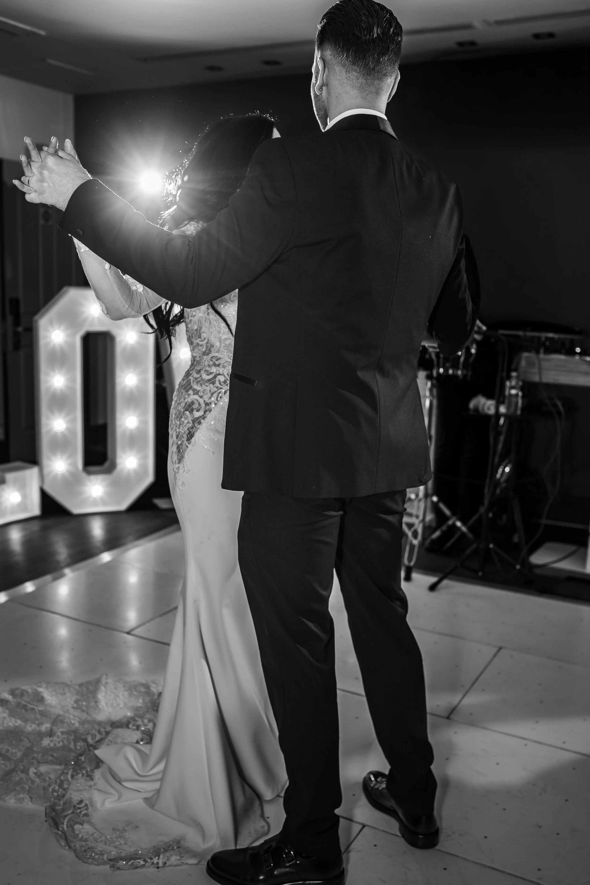 A couple dancing at a wedding reception, with the bride in a lace wedding dress and the groom in a dark suit, illuminated by a bright light and decorative illuminated letters in the background.