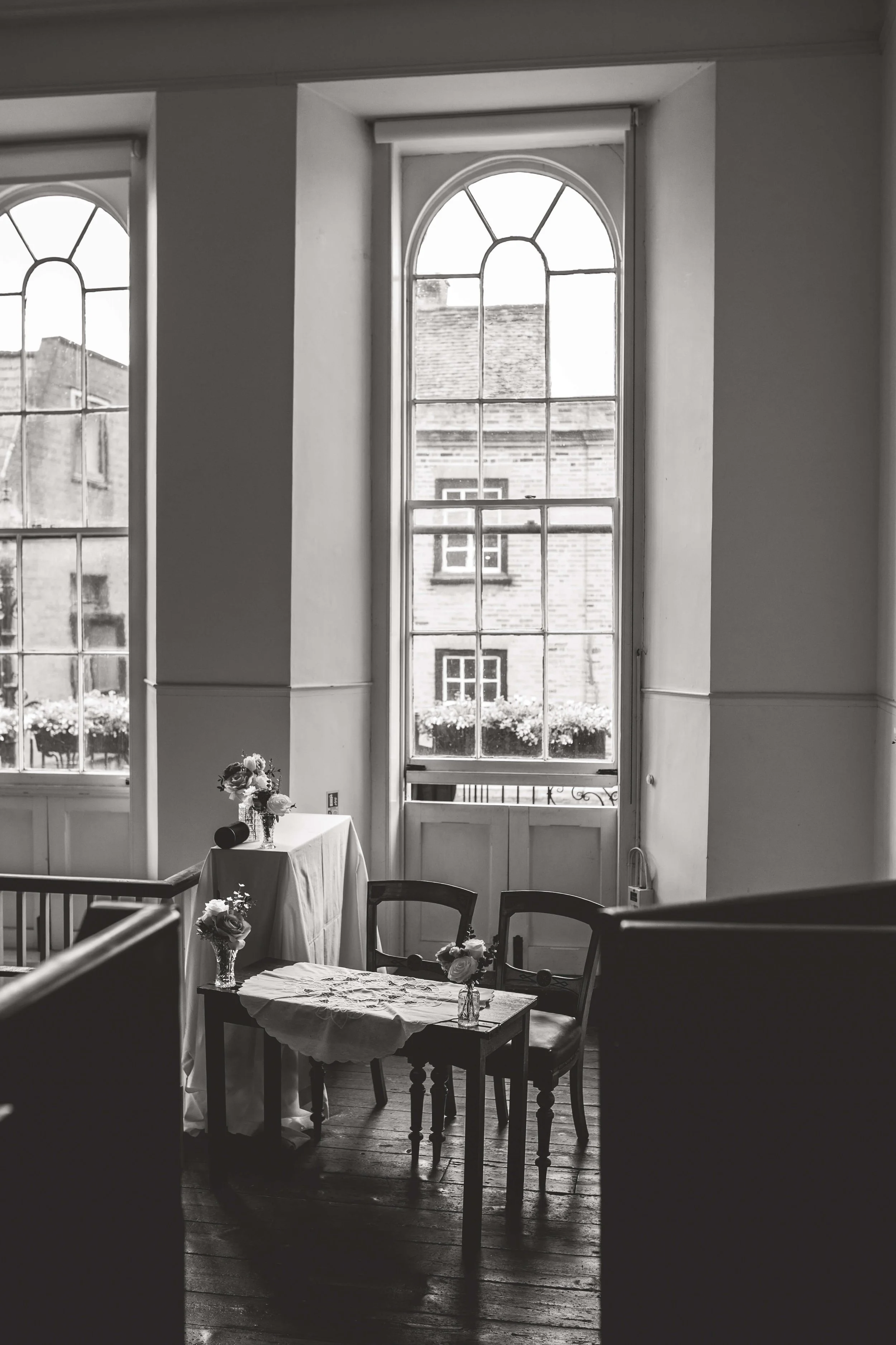 Interior of a room with large arched windows, small table with two chairs and floral arrangements, black-and-white photograph.