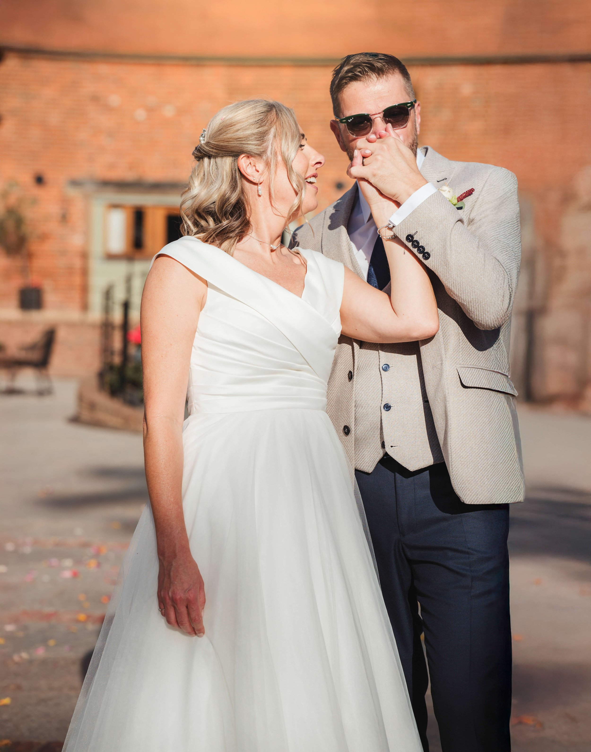A bride and groom sharing a joyful moment at their wedding, outside with a brick building in the background.