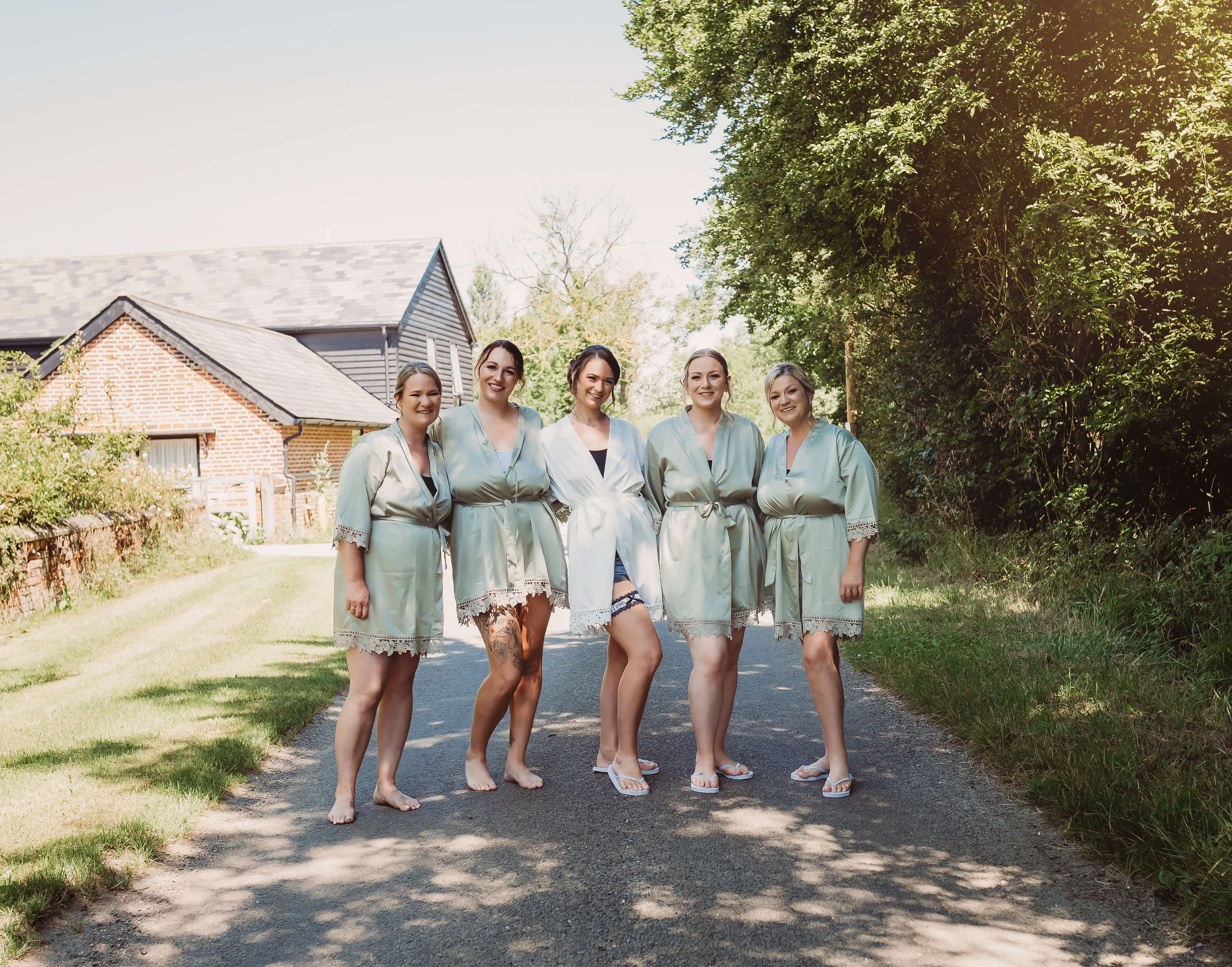 Group of five women wearing matching satin robes, smiling outdoors on a sunny day, standing on a paved path with houses and trees in the background.