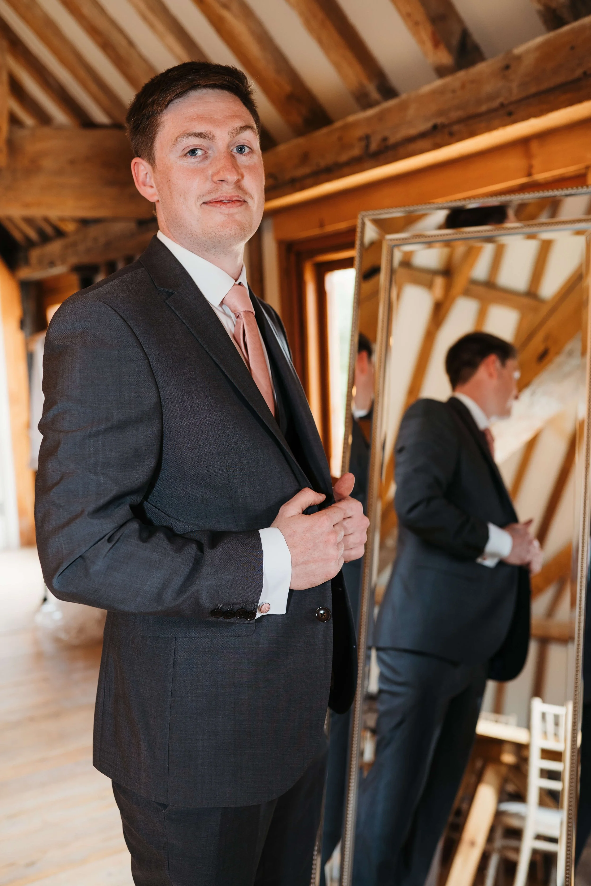 A man in a dark suit and pink tie looking at the camera while standing in front of a large mirror, adjusting his suit jacket, in a room with wooden beams and natural light.