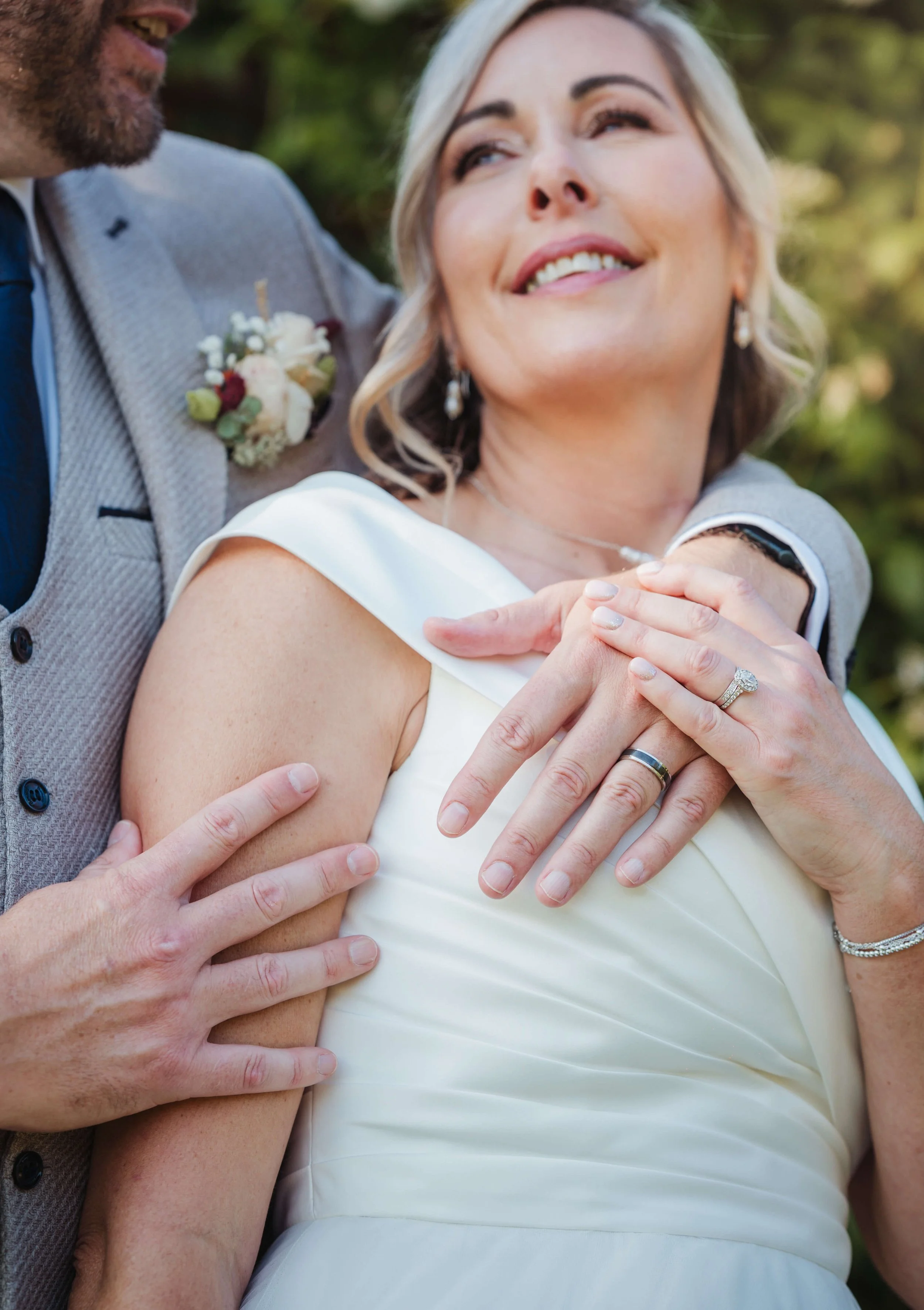 Close-up of a smiling woman wearing a white dress and jewelry, embracing and being embraced by a man in a gray suit with a boutonniere, outdoors with a blurred green background.