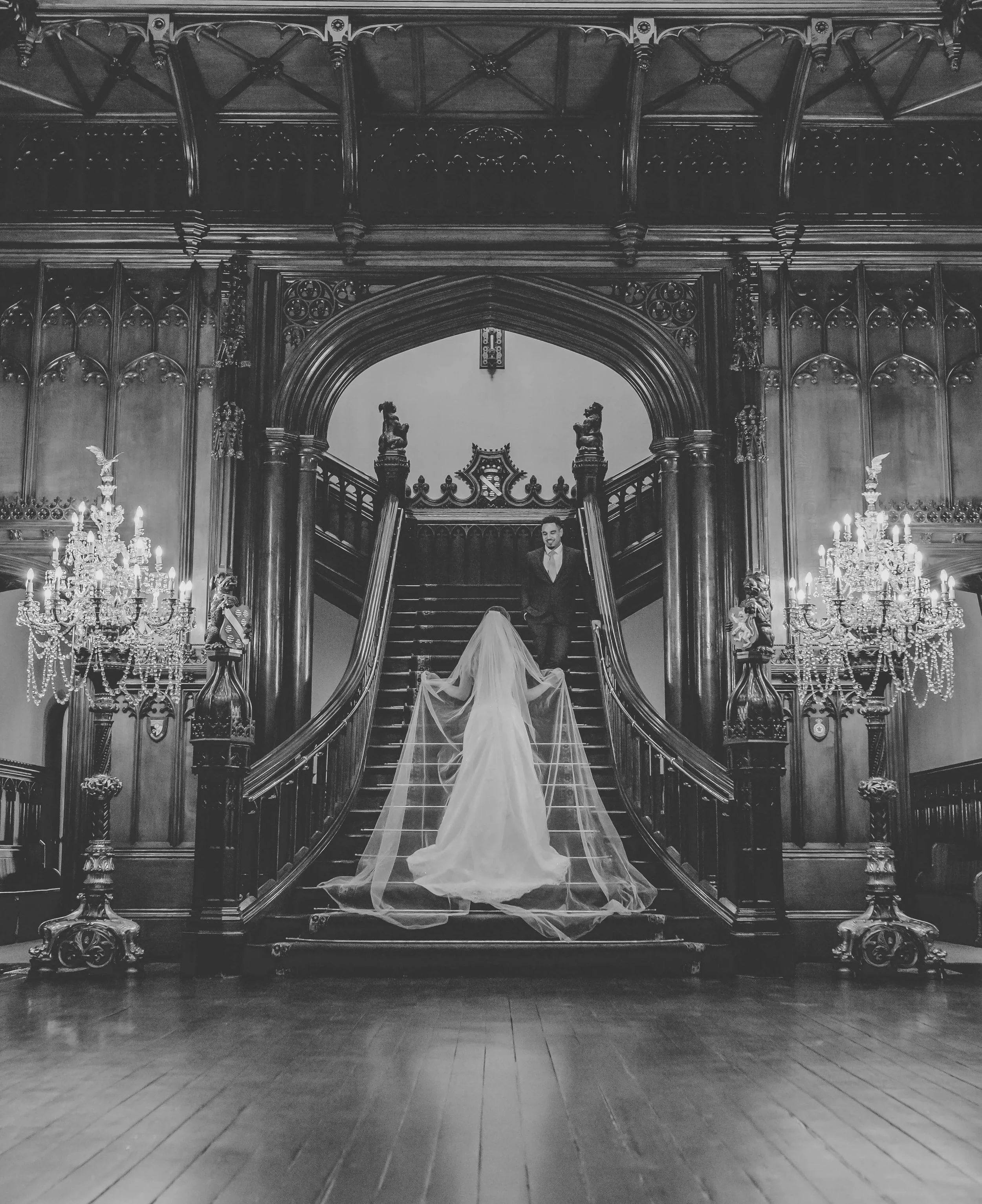 A bride in a long wedding dress and veil standing at the bottom of a grand wooden staircase, facing a groom in a suit standing at the top, in an ornate, luxurious interior with chandeliers and intricate woodwork.