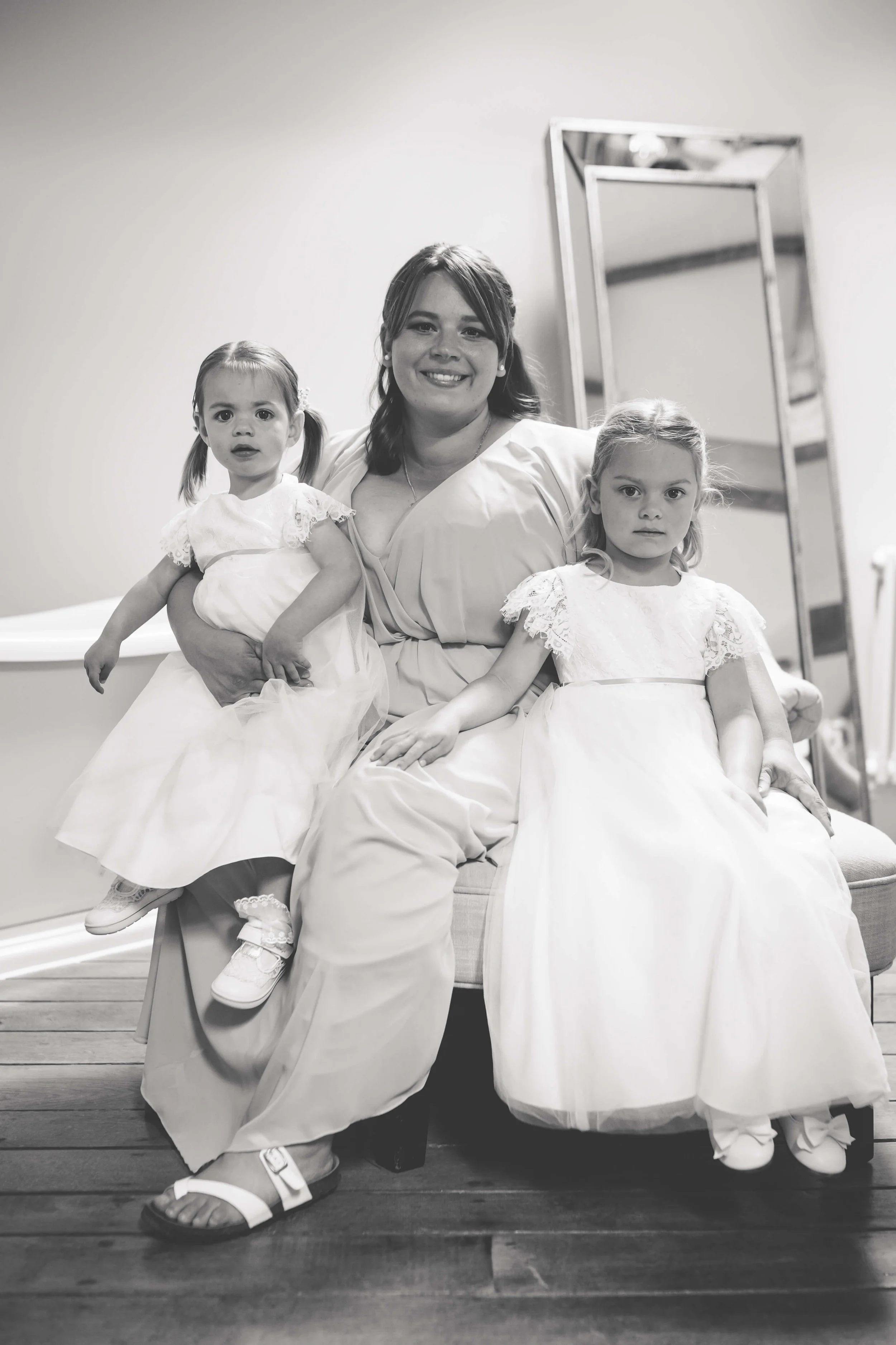A woman sitting on a couch with two young girls, all dressed in white, in a room with a mirror in the background.