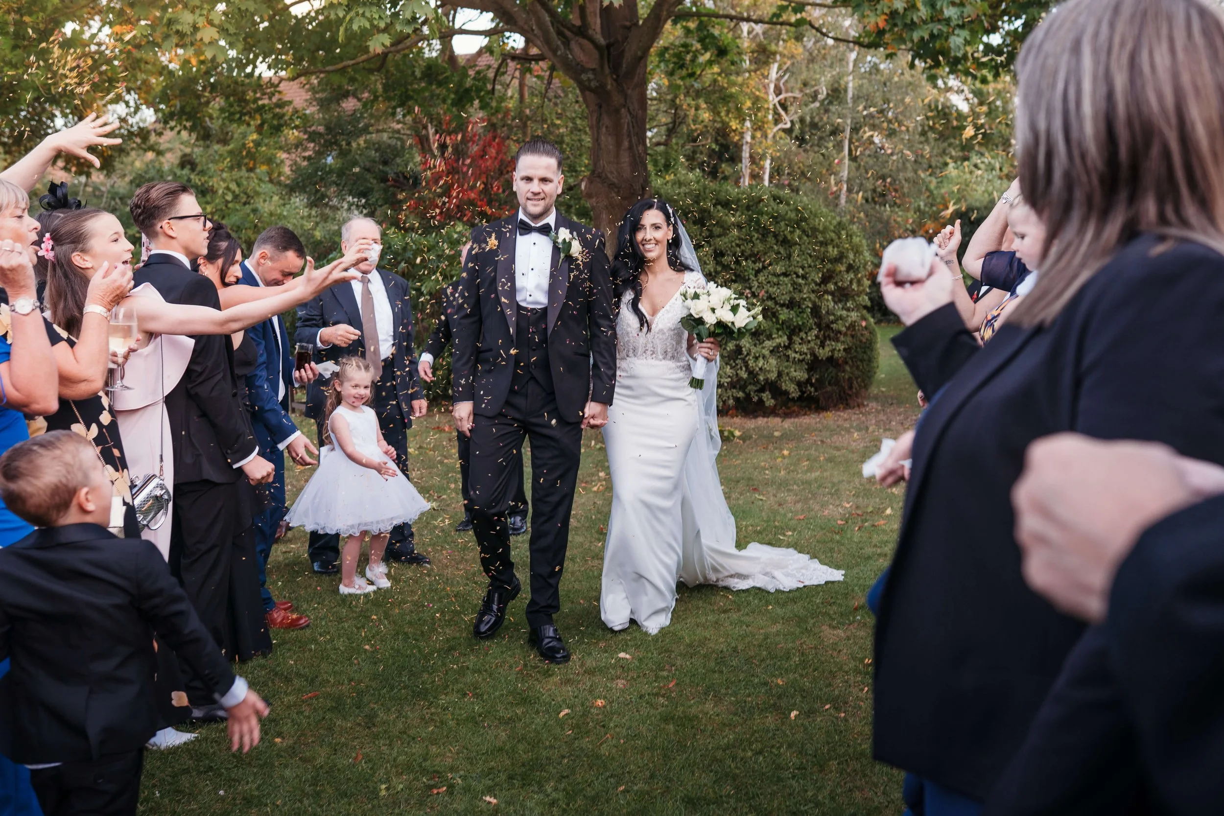A newlywed couple walks hand in hand through a crowd of celebrating guests outdoors, with the bride holding a bouquet and the groom in a tuxedo. Confetti is falling, and trees with green and autumn-colored leaves are in the background.
