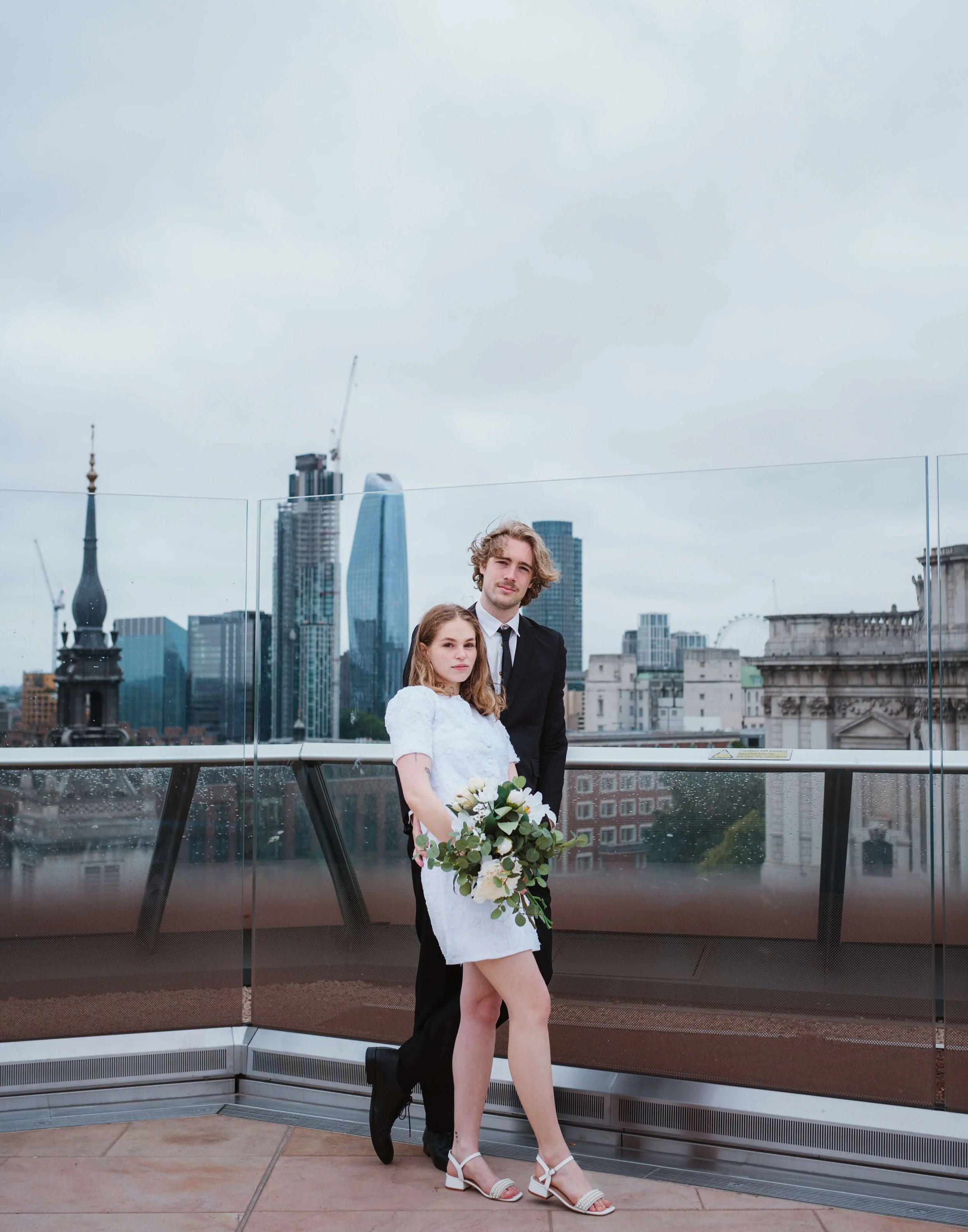 A young woman in a white dress and high heels holding a bouquet of flowers, standing in front of a young man in a black suit on a rooftop with a city skyline background.