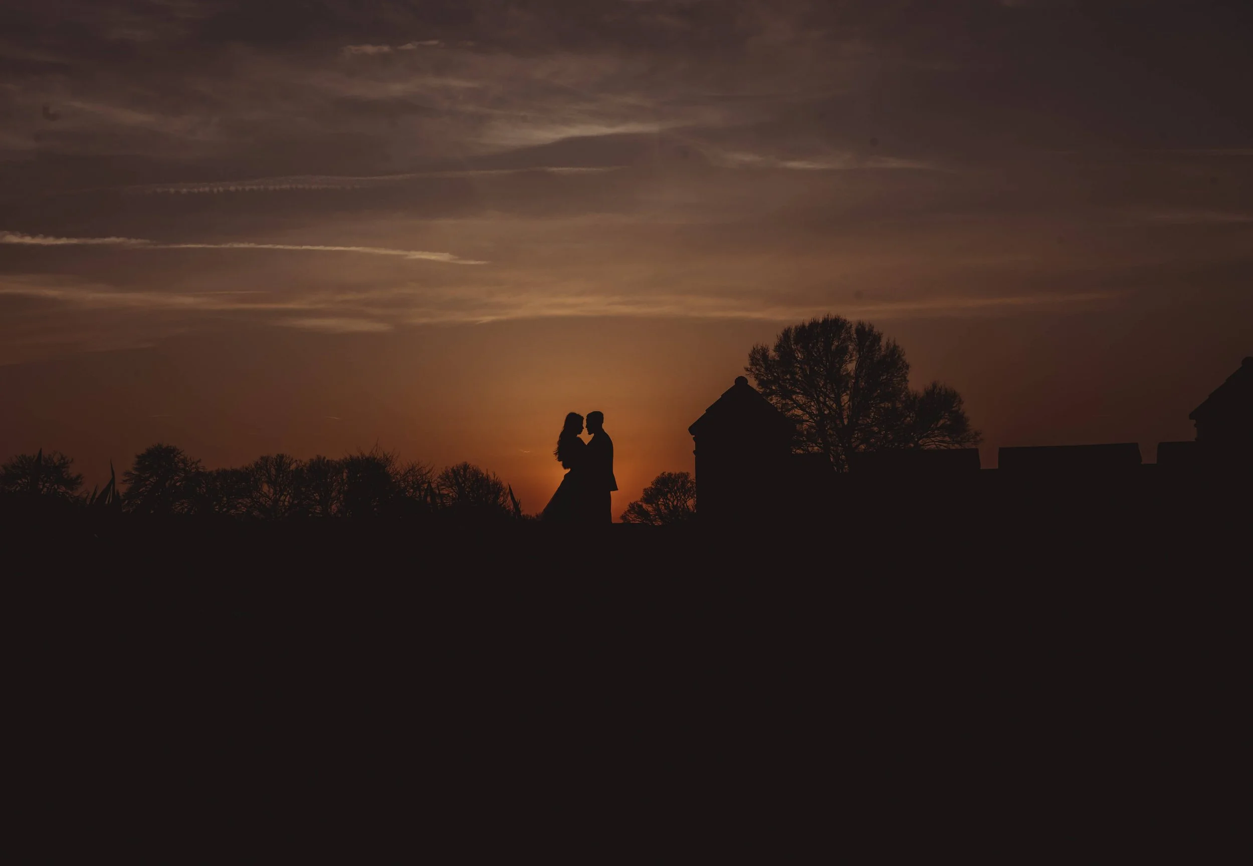 Silhouette of a couple embracing at sunset with trees and buildings in the background.