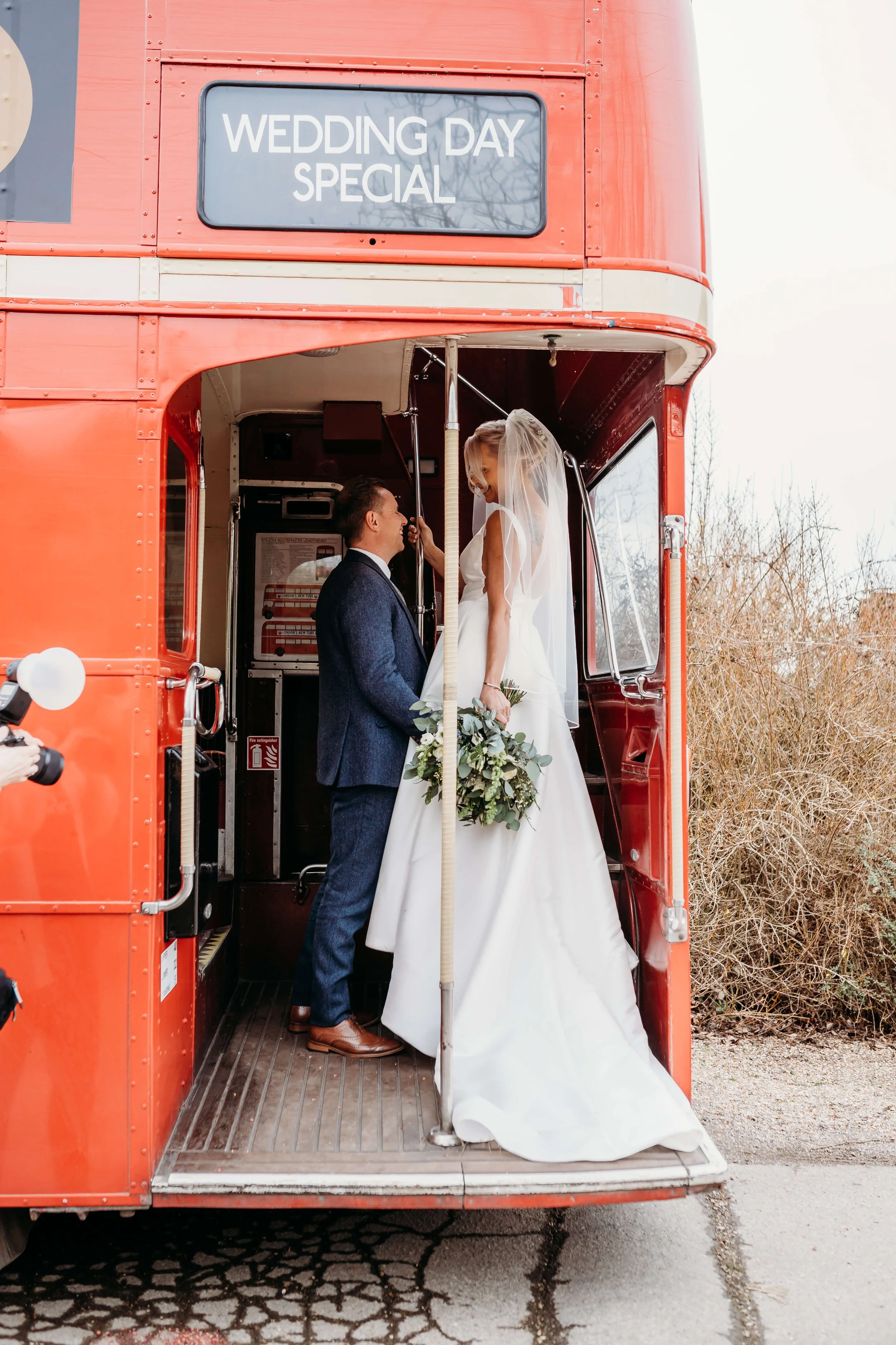 Bride and groom inside a red double-decker bus with a sign that reads 'Wedding Day Special.' The bride is standing on the bus's step, holding a bouquet of flowers, while the groom looks up at her.