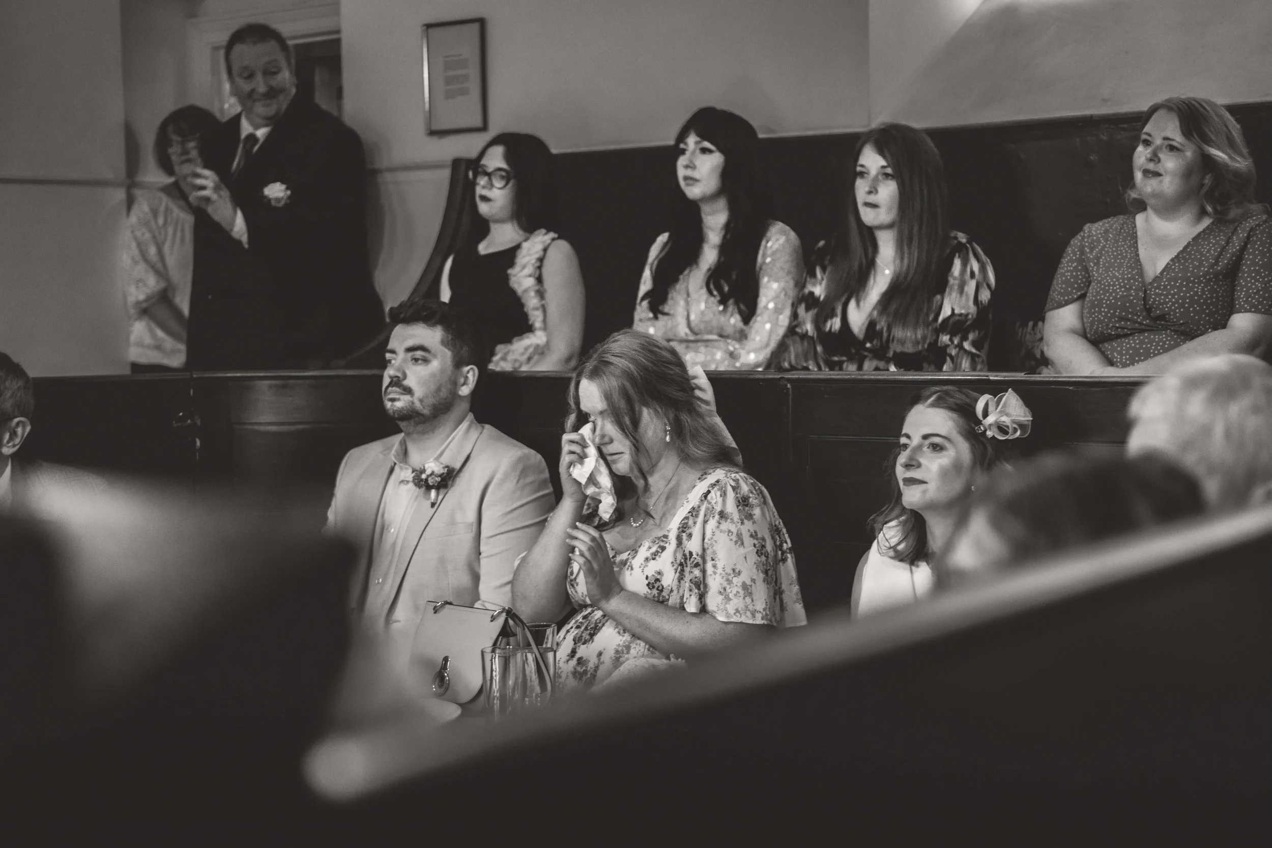 People seated at a formal event, some looking emotional and tearful, with a man in a suit on the left smiling and an older woman wiping her tears.