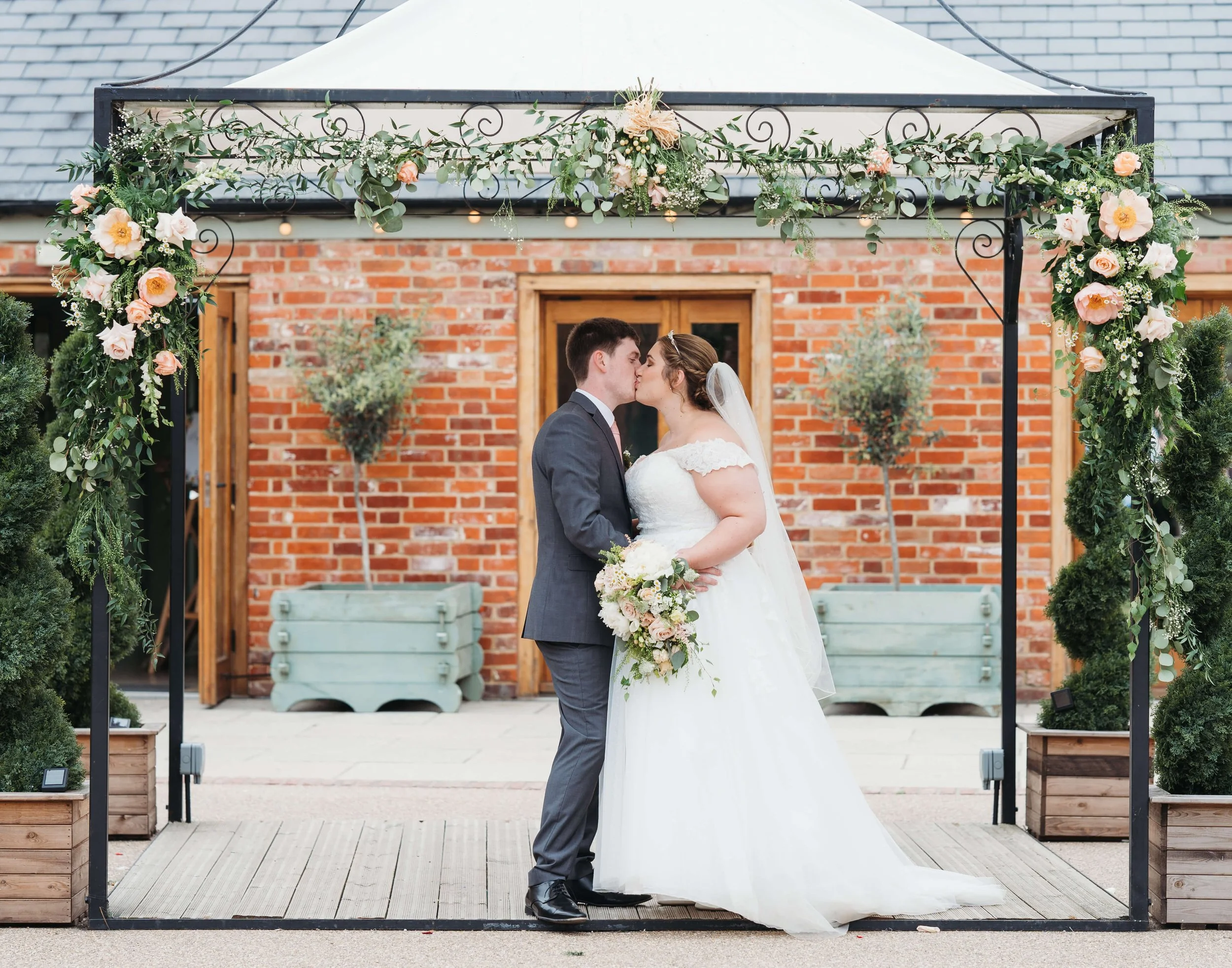 A bride and groom share a kiss during their wedding ceremony under a floral archway. The bride wears a white wedding gown and holds a bouquet, while the groom is dressed in a gray suit. The setting features a brick wall background and potted plants.
