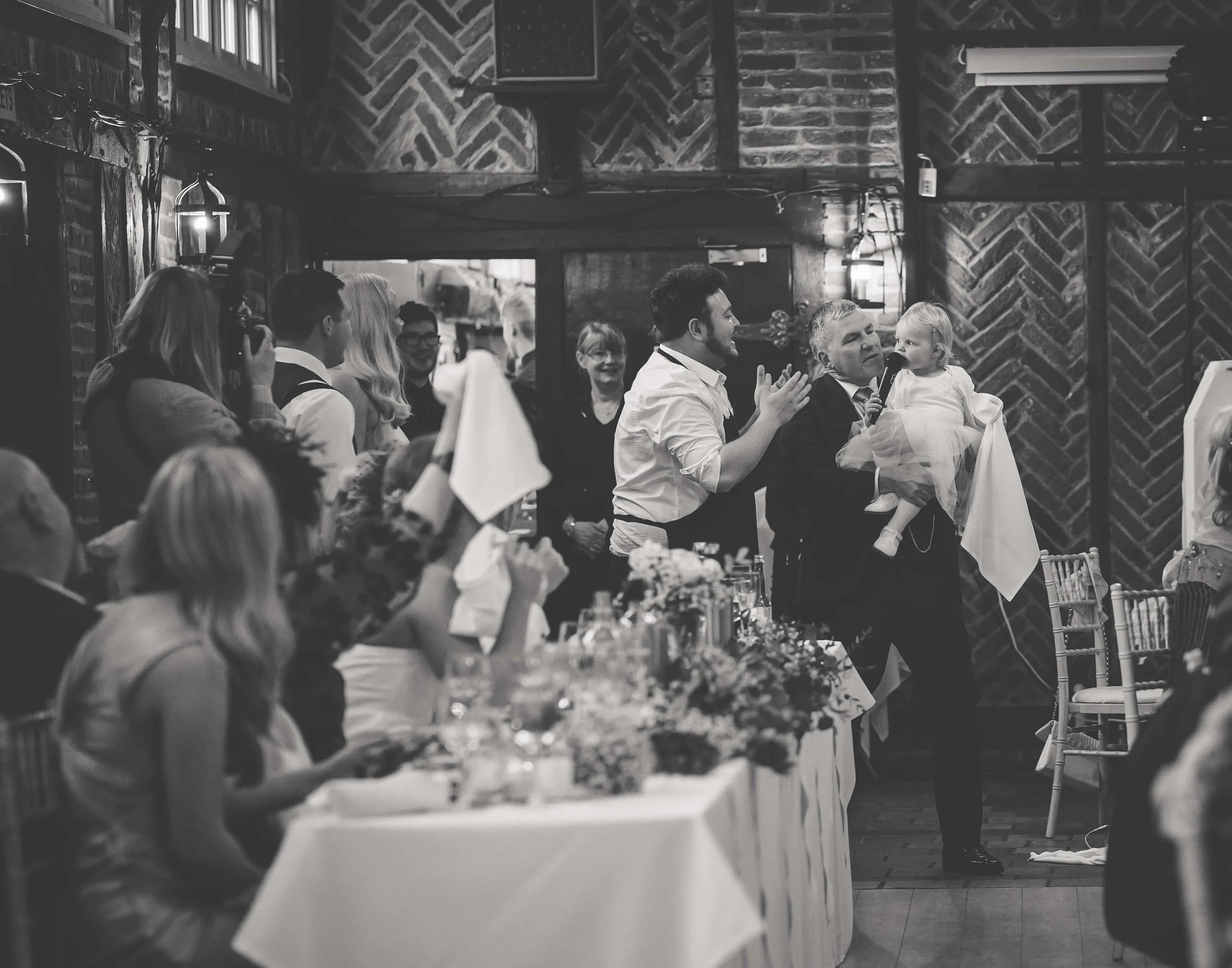 People gathered at a celebration, possibly a wedding, with some raising glasses. A man is holding a young girl, while a woman is clapping nearby. Guests are seated at tables with floral arrangements, and the setting has a rustic interior with brick w