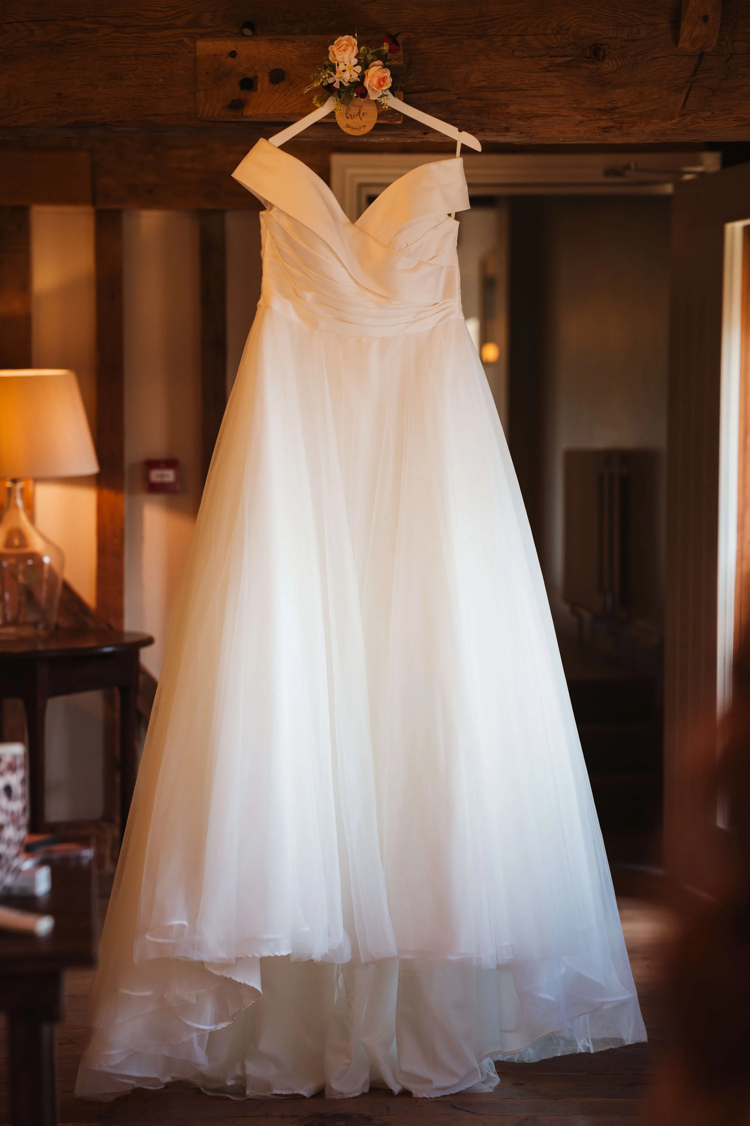 Elegant white wedding dress hanging on a floral hanger in a warmly lit rustic room.