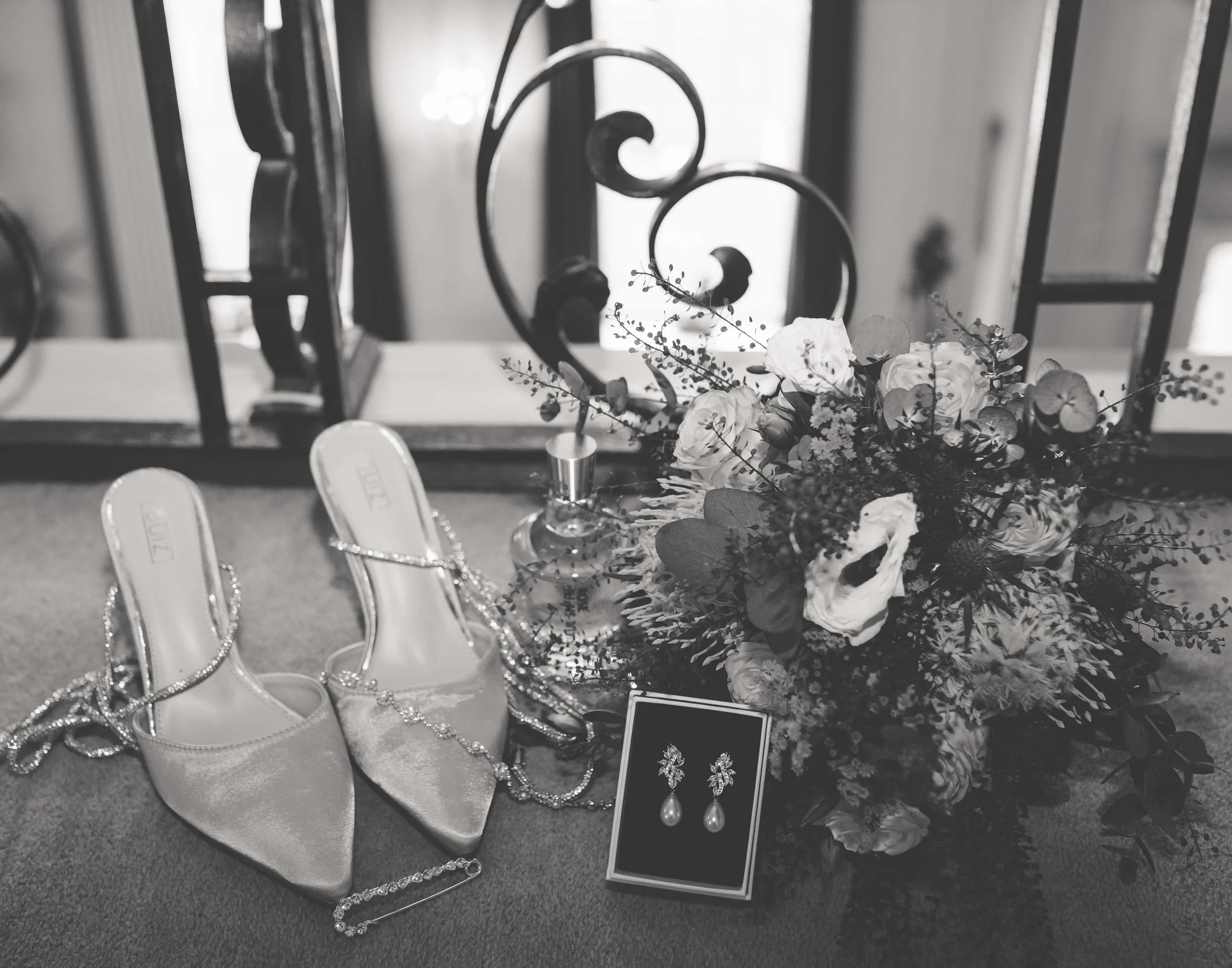 Black and white photo of a table with a bouquet of flowers, a pair of high-heeled shoes, pearl jewelry, earrings, and a perfume bottle, set before a decorative wrought iron railing.