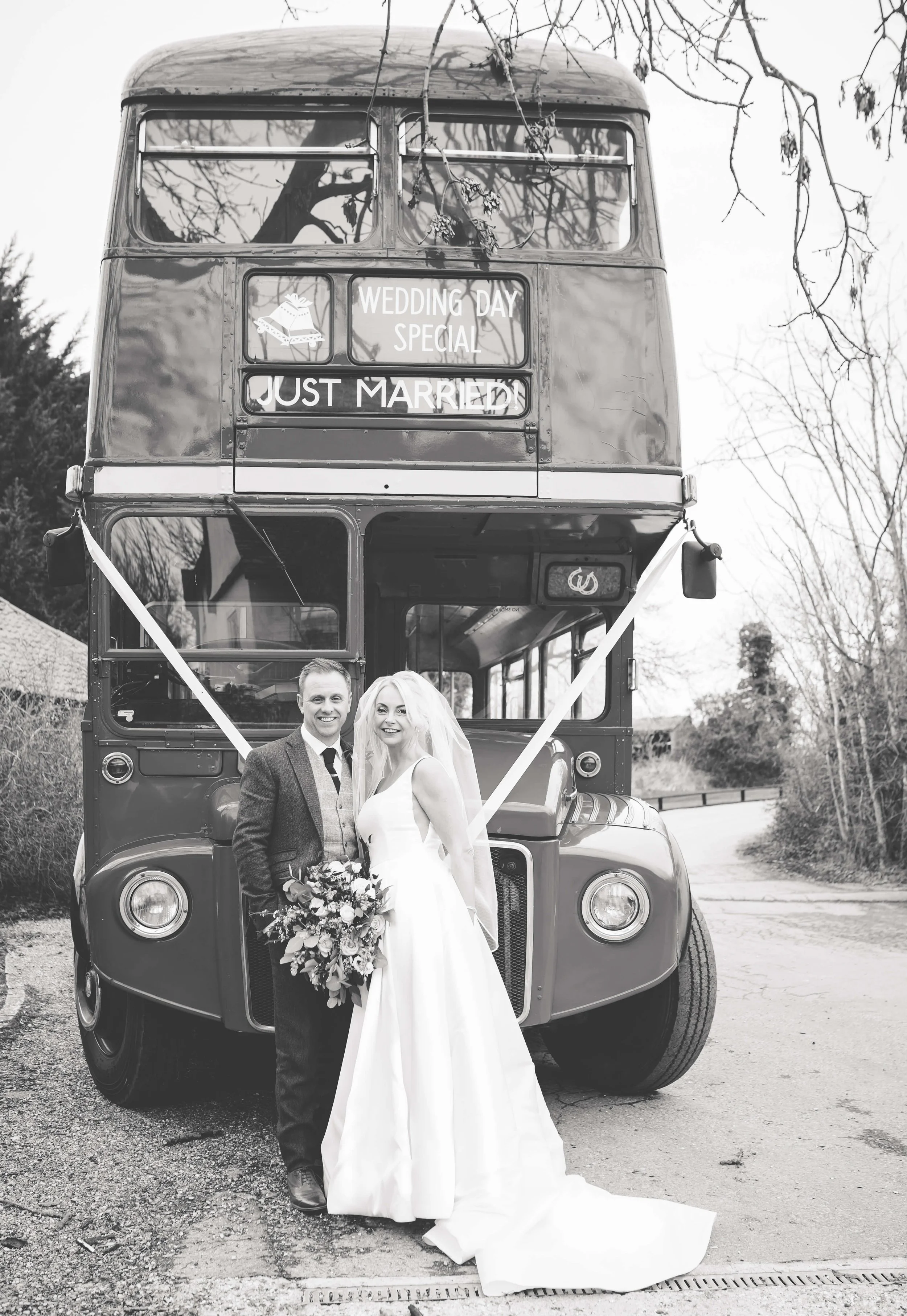 A newly married couple in wedding attire standing in front of a vintage double-decker bus decorated with ribbons. The bus has a sign that reads, "Wedding Day Special Just Married." The couple is smiling, and the bride is holding a bouquet of flowers.