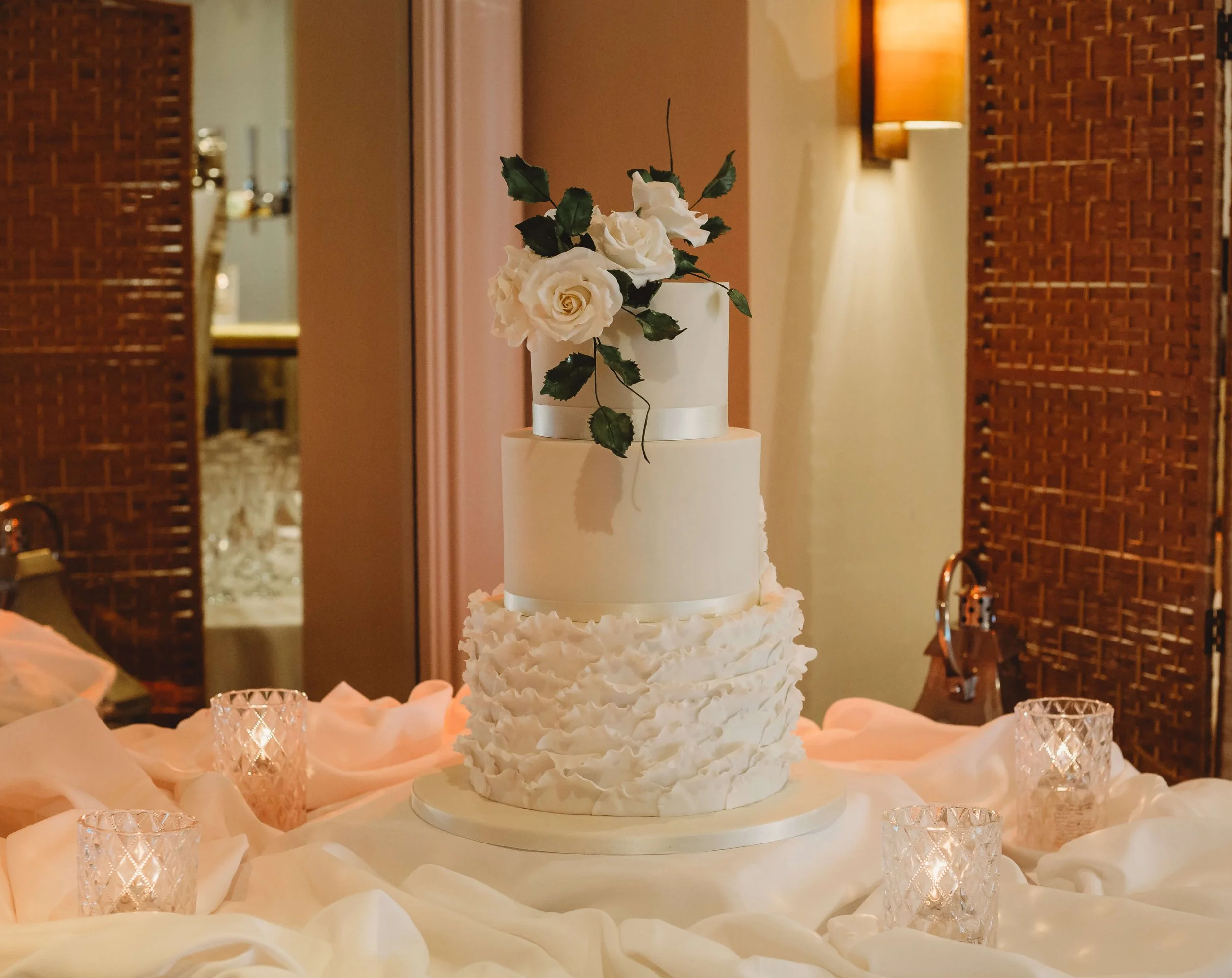 Elegant white wedding cake with three tiers, decorated with white roses and greenery on top, on a table with soft pink and white fabric and four glass candle holders surrounding it.