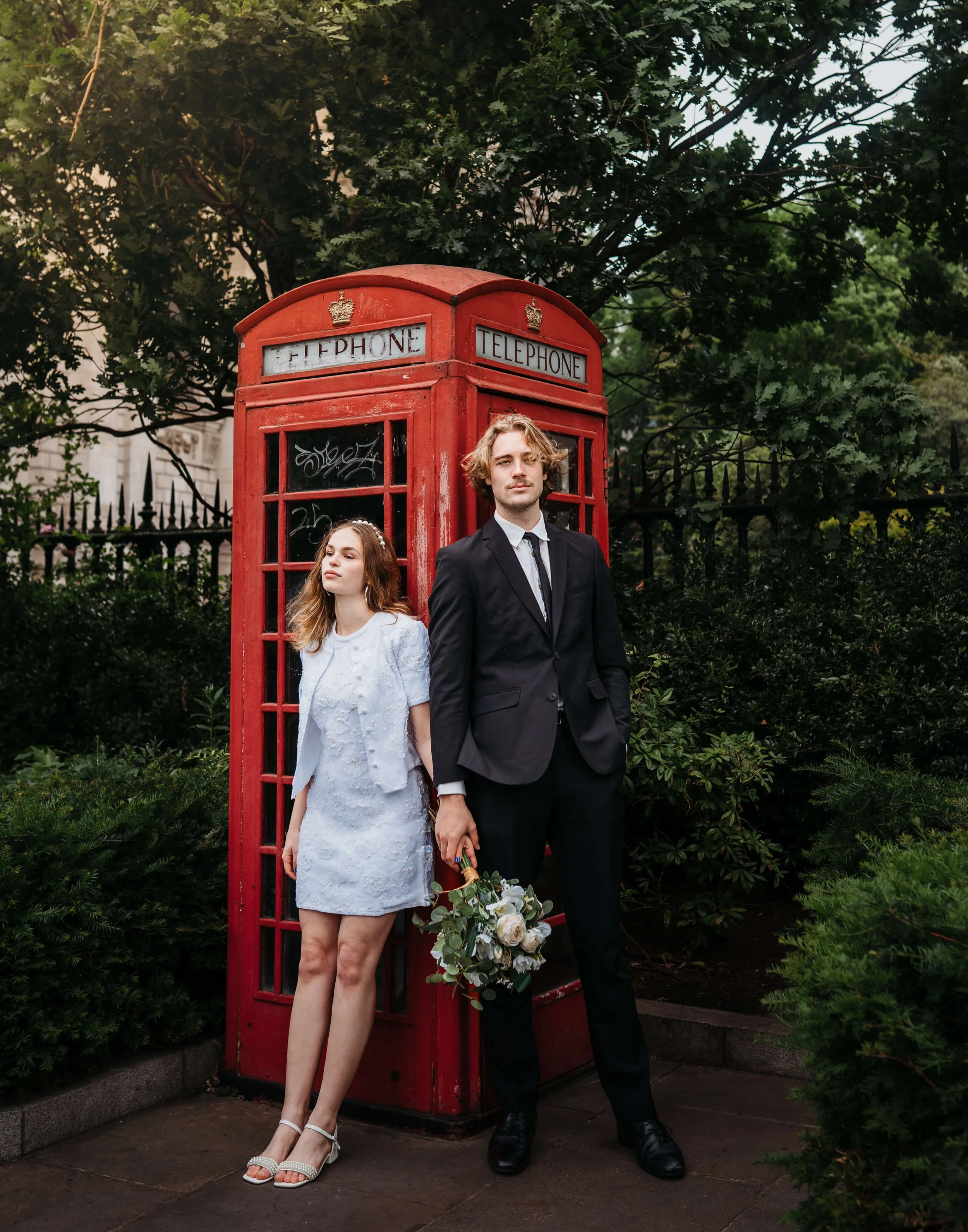 A young couple standing next to a classic red telephone booth outdoors, with green trees and foliage in the background. The woman is wearing a white dress and heels, and the man is wearing a black suit and holding a bouquet of flowers.