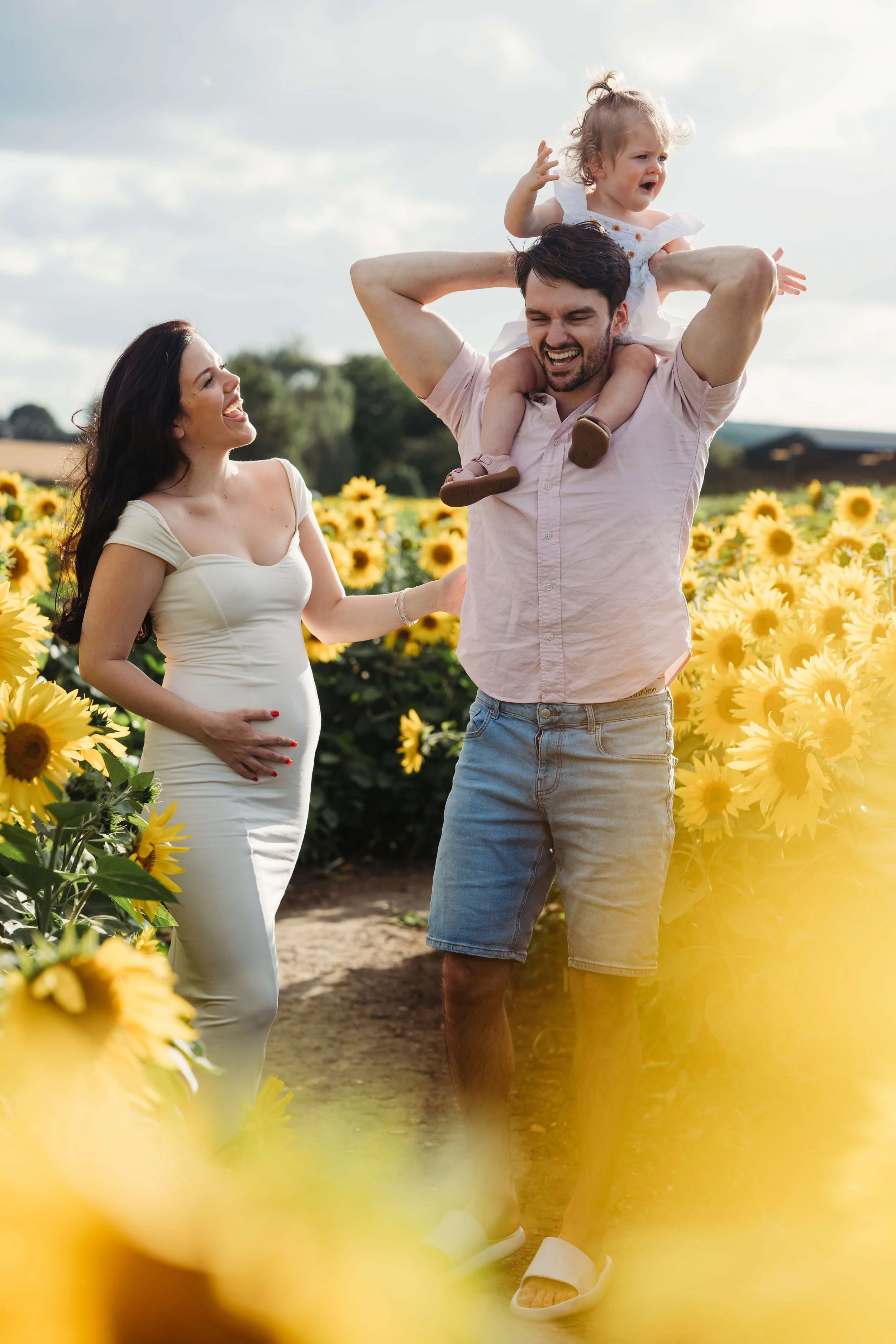 A family of three enjoying a sunny day in a sunflower field. The man is carrying a young girl on his shoulders, and the woman, who appears pregnant, is smiling at them.