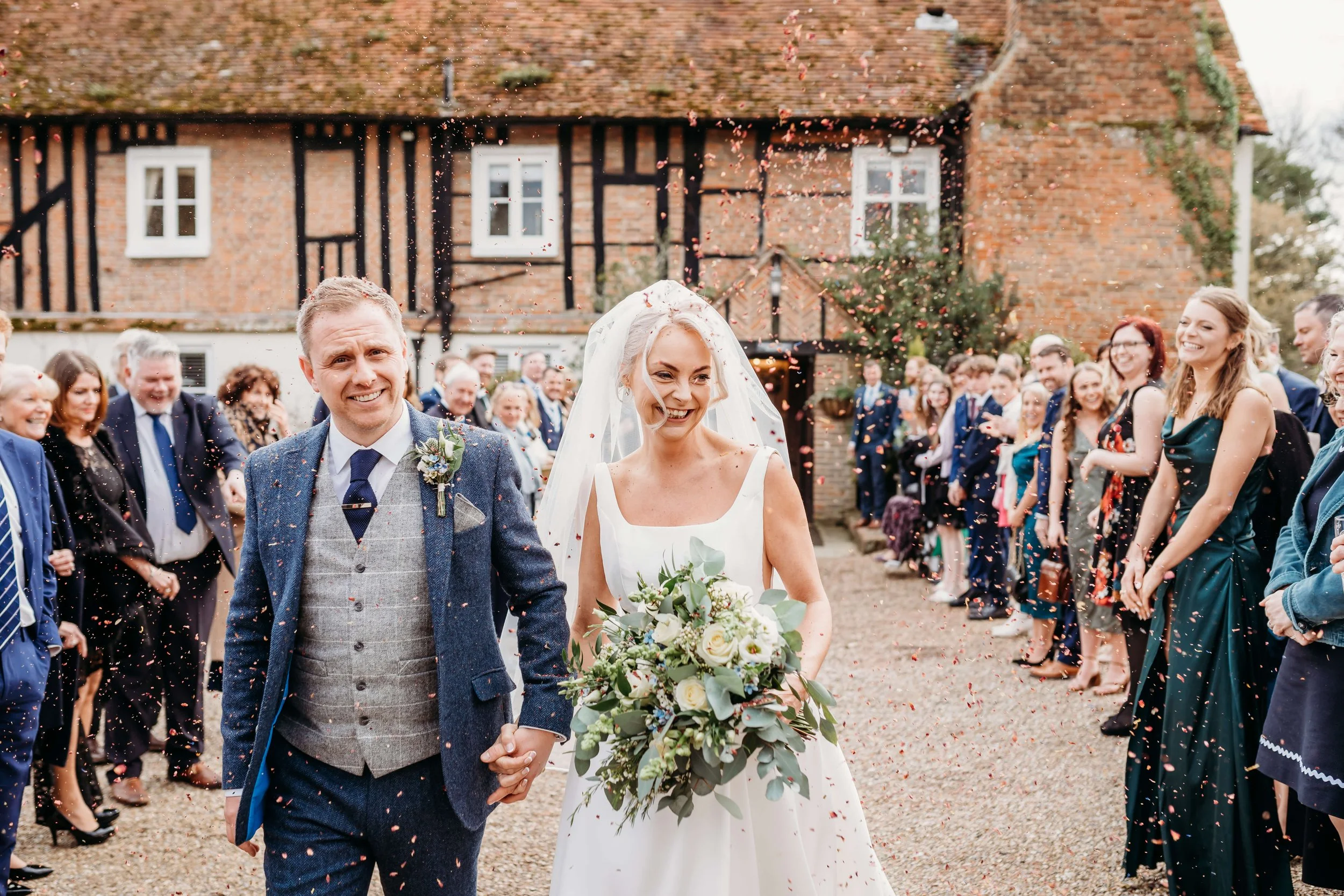 A bride and groom walking hand in hand, smiling, as guests celebrate their wedding before a rustic house, with confetti falling around them.