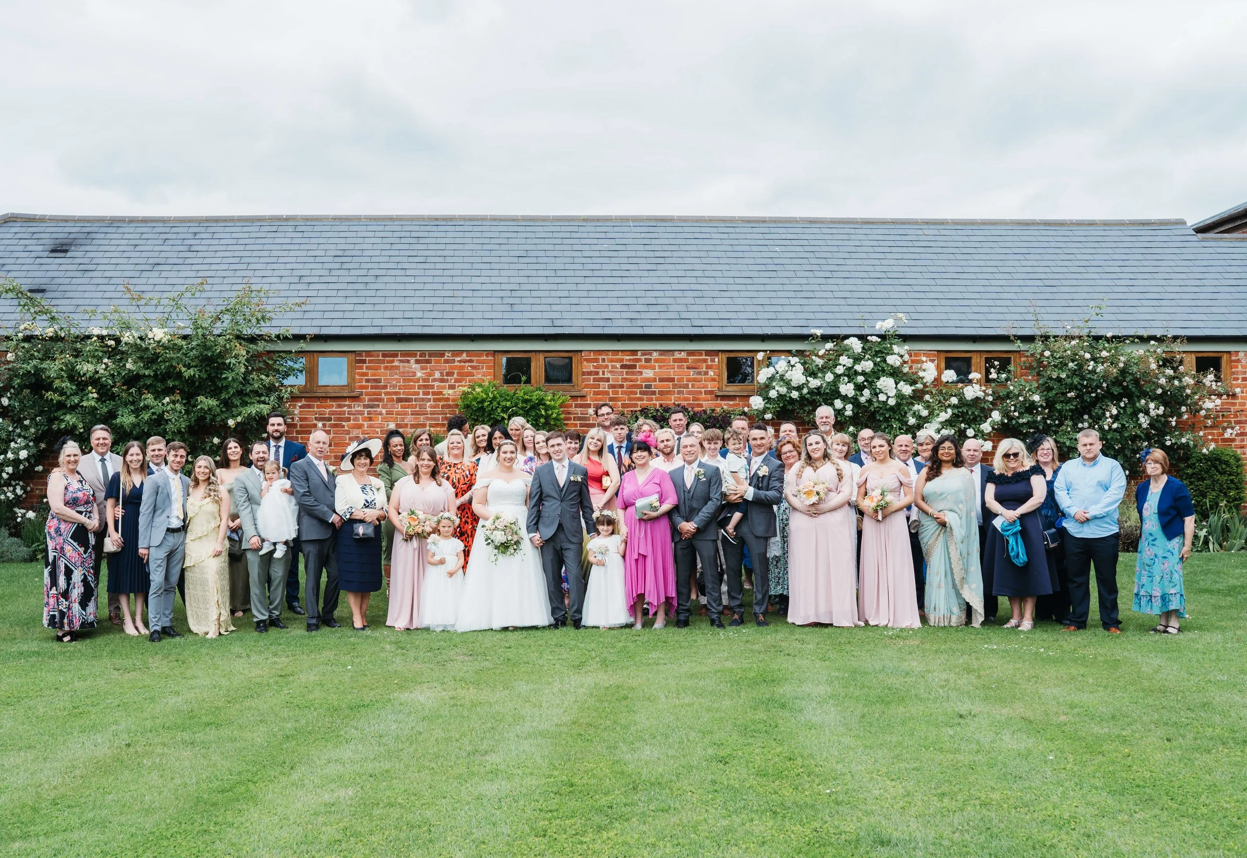 Large wedding party standing on a lawn with a brick building and flowering bushes in the background.