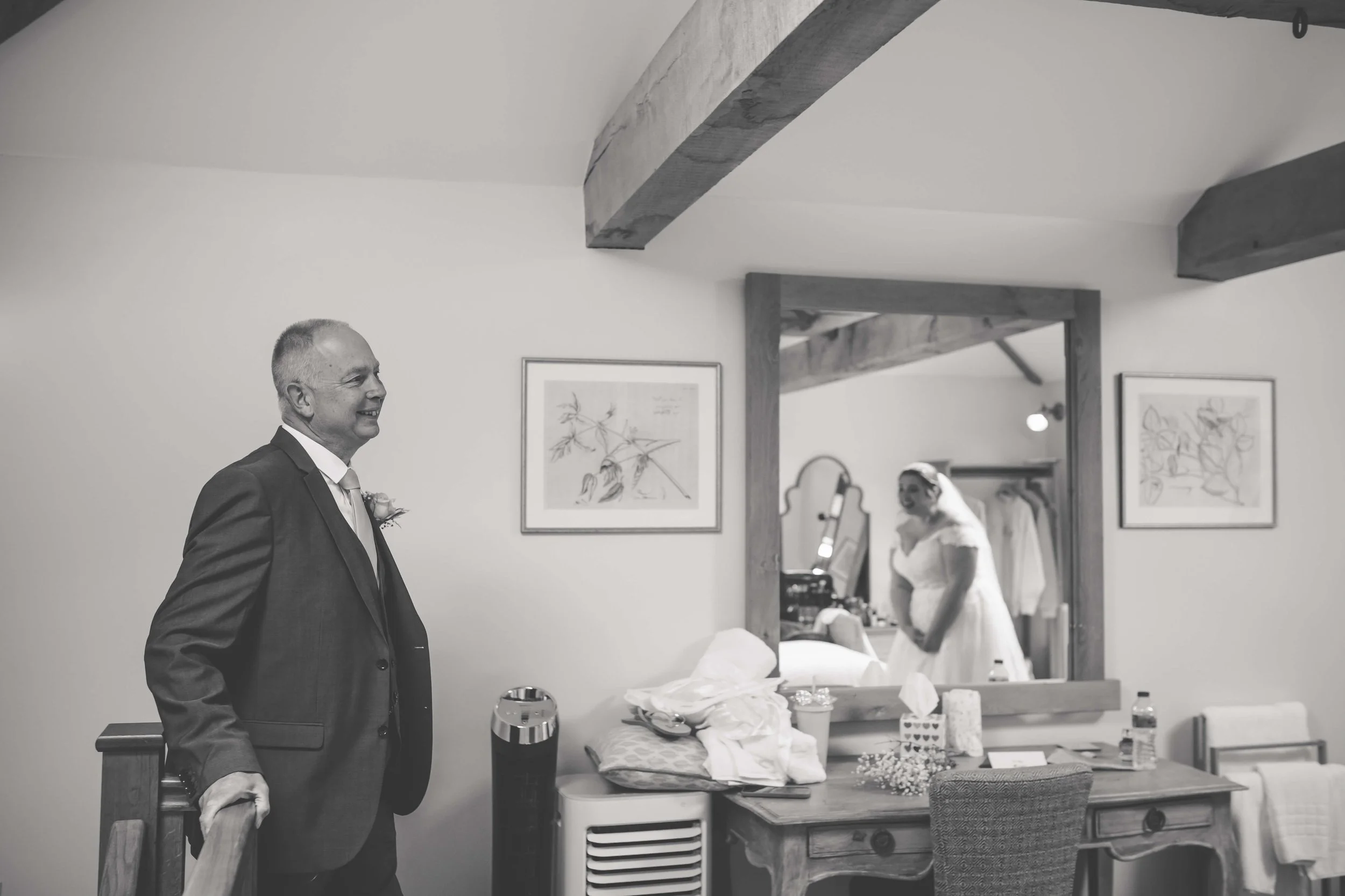 A man dressed in a suit stands smiling in a room decorated with framed artwork, with a bride visible in the mirror behind him, preparing for a wedding.