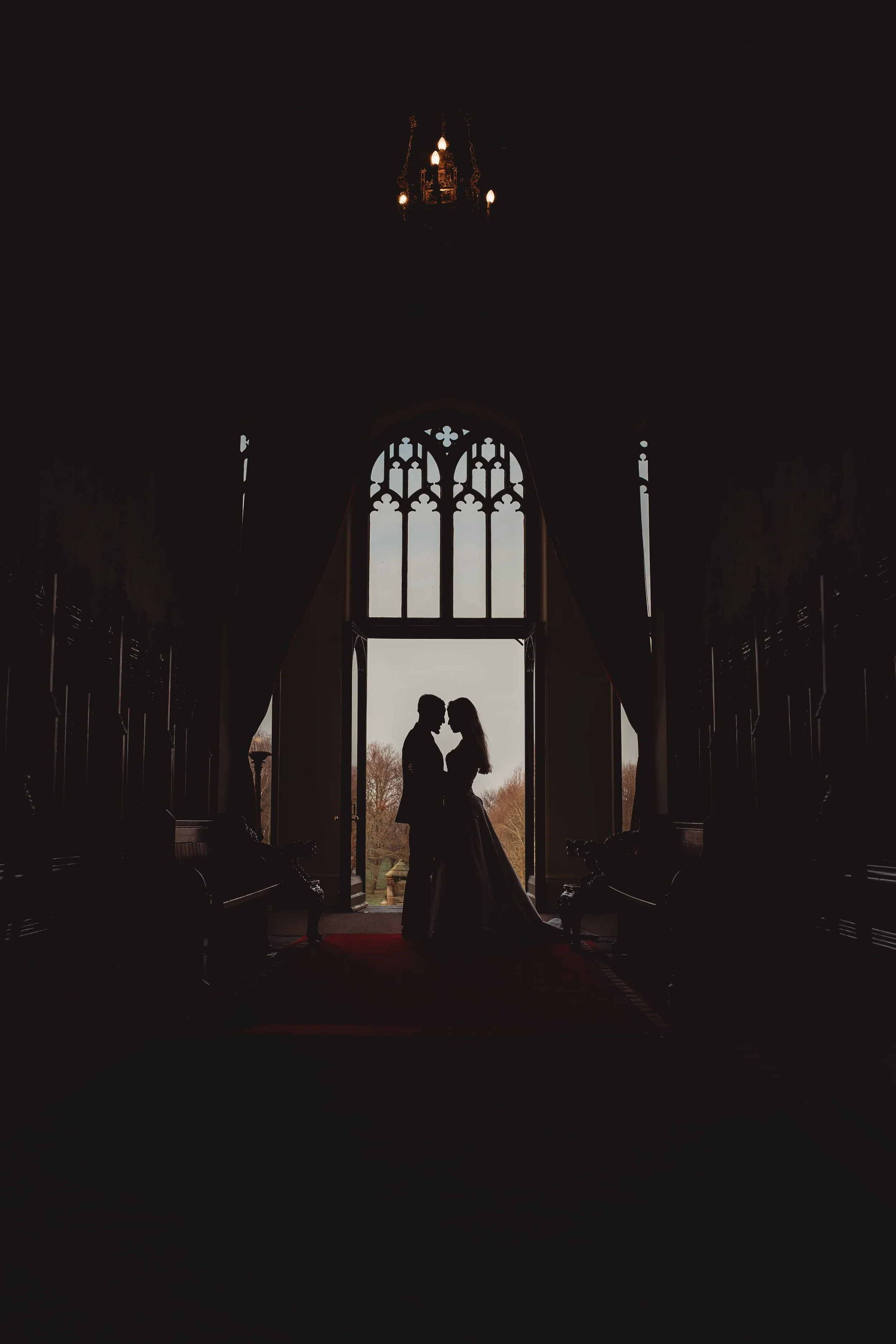 Silhouette of a couple standing close together, looking at each other inside a gothic-style church or castle with large stained glass windows, illuminated by natural light from outside.