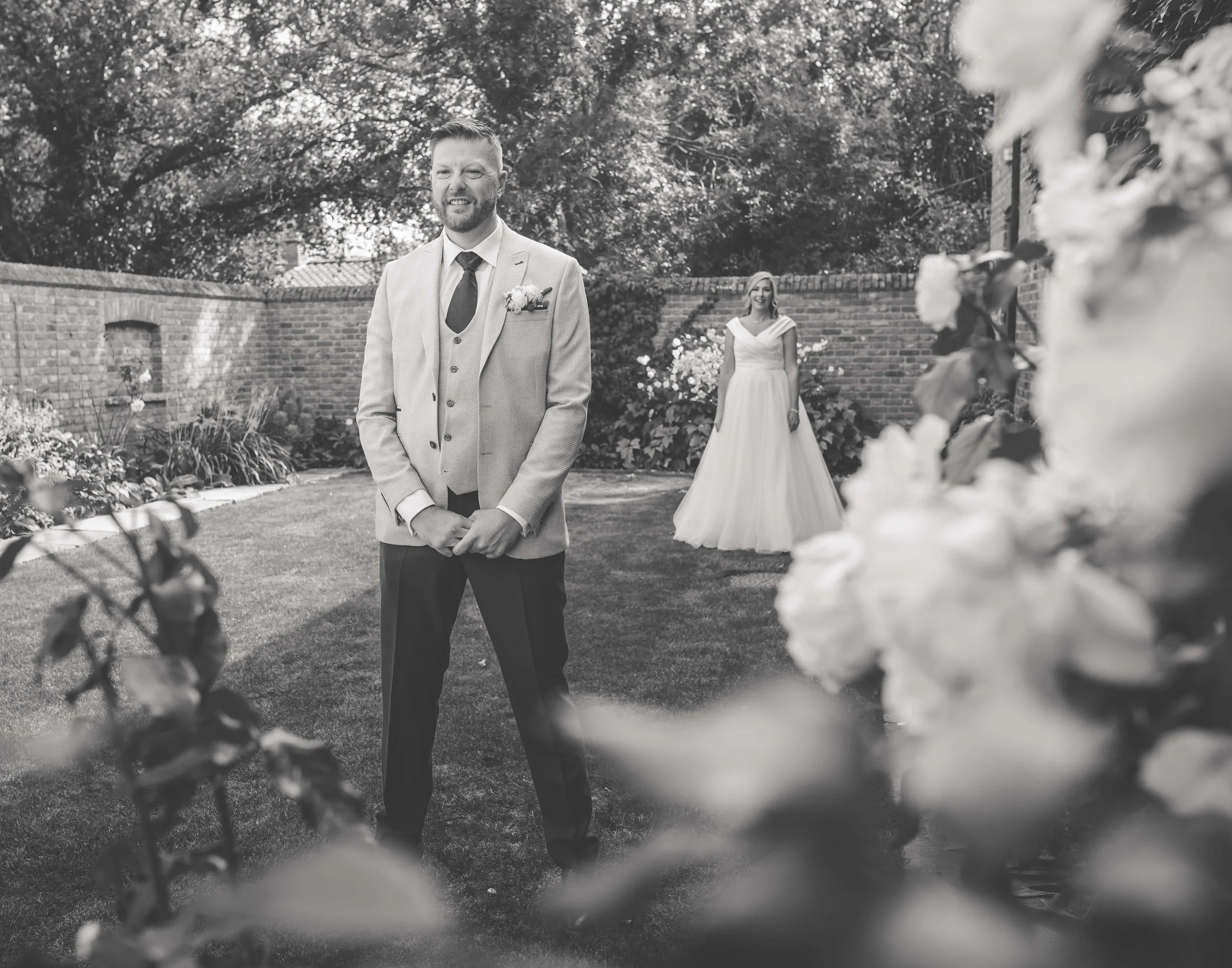 Black and white photo of a groom standing and smiling in a wedding garden, with a bride in a wedding dress in the background, surrounded by flowers and trees.