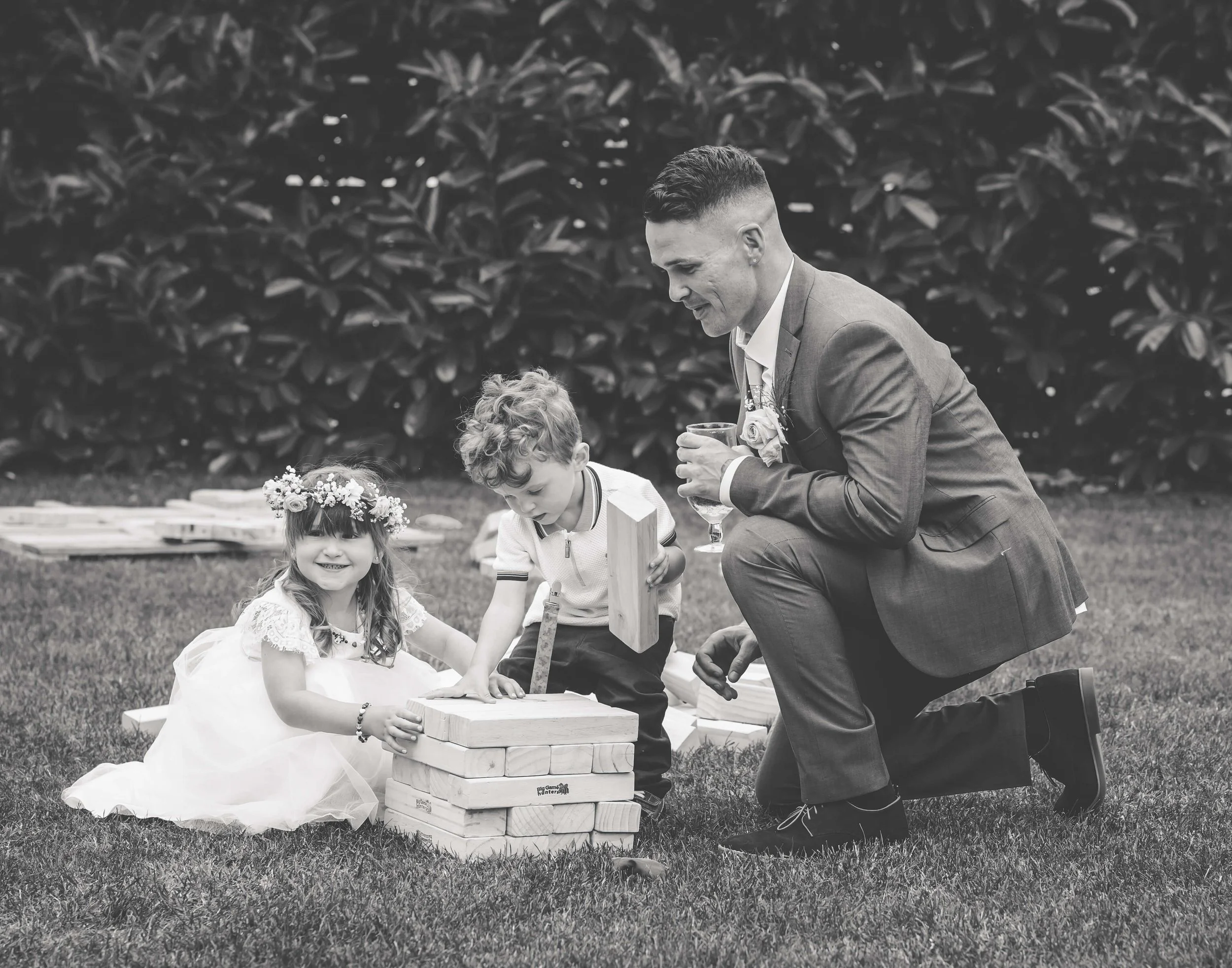 A man in a suit kneeling on grass, holding a glass, and looking at two young children playing a giant Jenga game outdoors. The girl wears a white dress with a flower crown, and the boy wears a light-colored shirt. Both children are focused on the gam