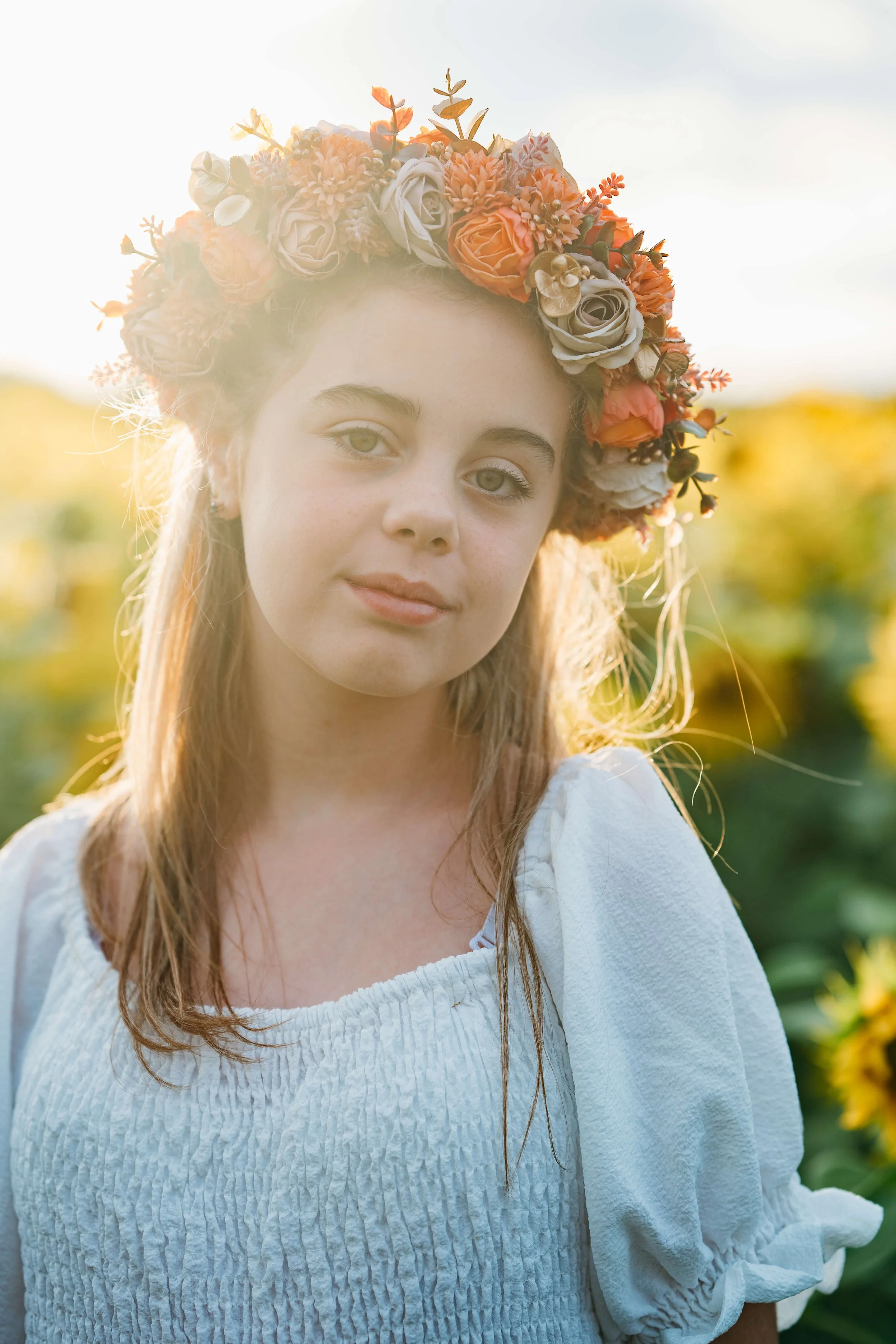 A young woman with light-colored hair wearing a colorful flower crown and a white dress, standing outdoors in sunlight, with a field of sunflowers in the background.