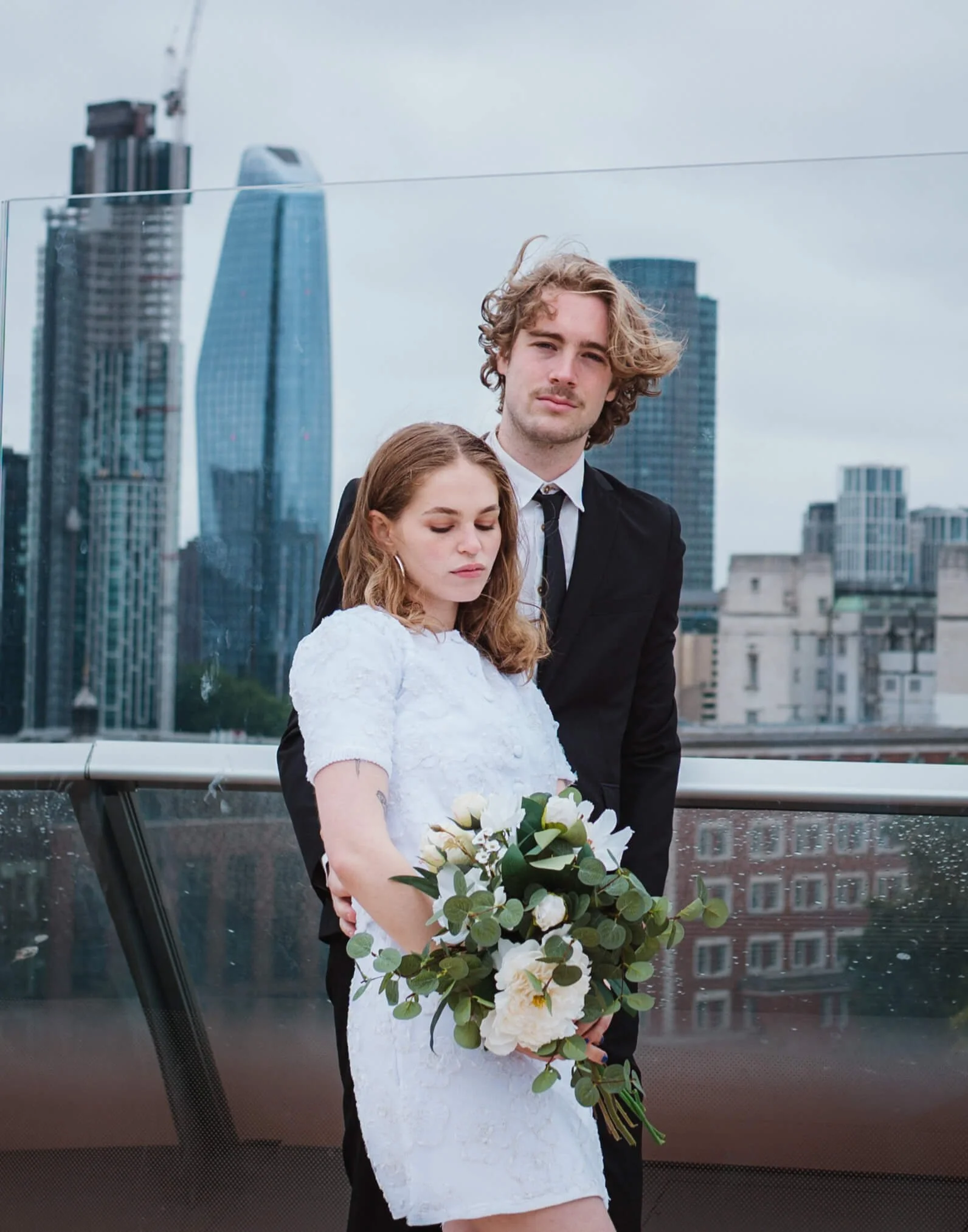 A young couple dressed in wedding attire, standing on a rooftop with a city skyline in the background. The woman is holding a bouquet of white flowers and greenery, with her eyes closed. The man is standing behind her, looking at the camera with a ne