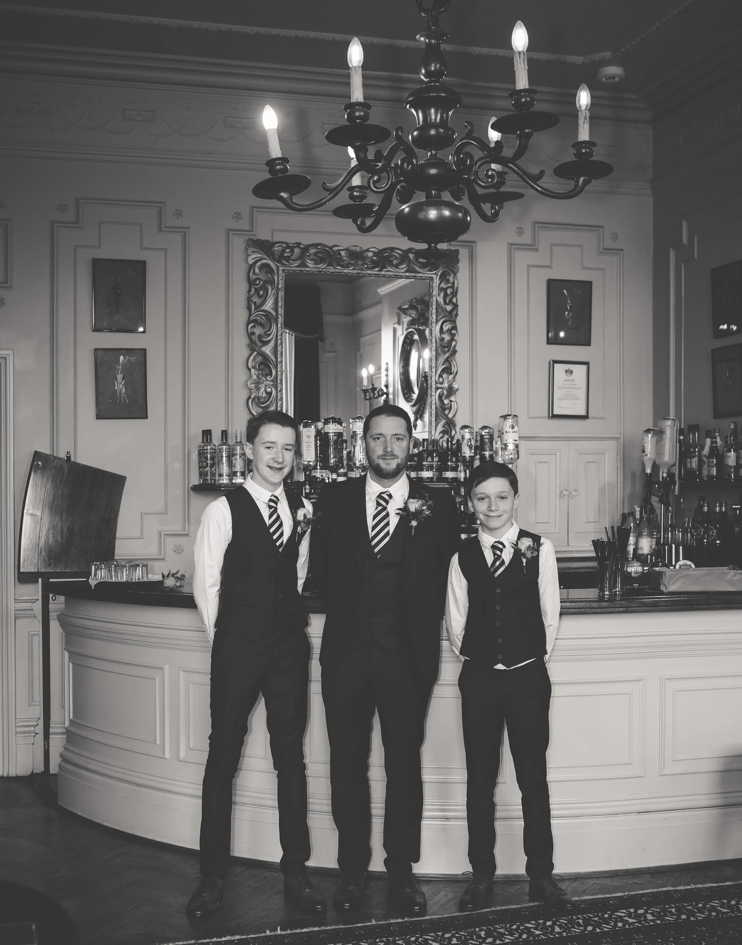 Black-and-white photo of three boys dressed in formal vests, shirts, and striped ties, standing in front of a fancy bar with a chandelier above. The boy in the middle has a beard, the other two are younger with short hair, all smiling.