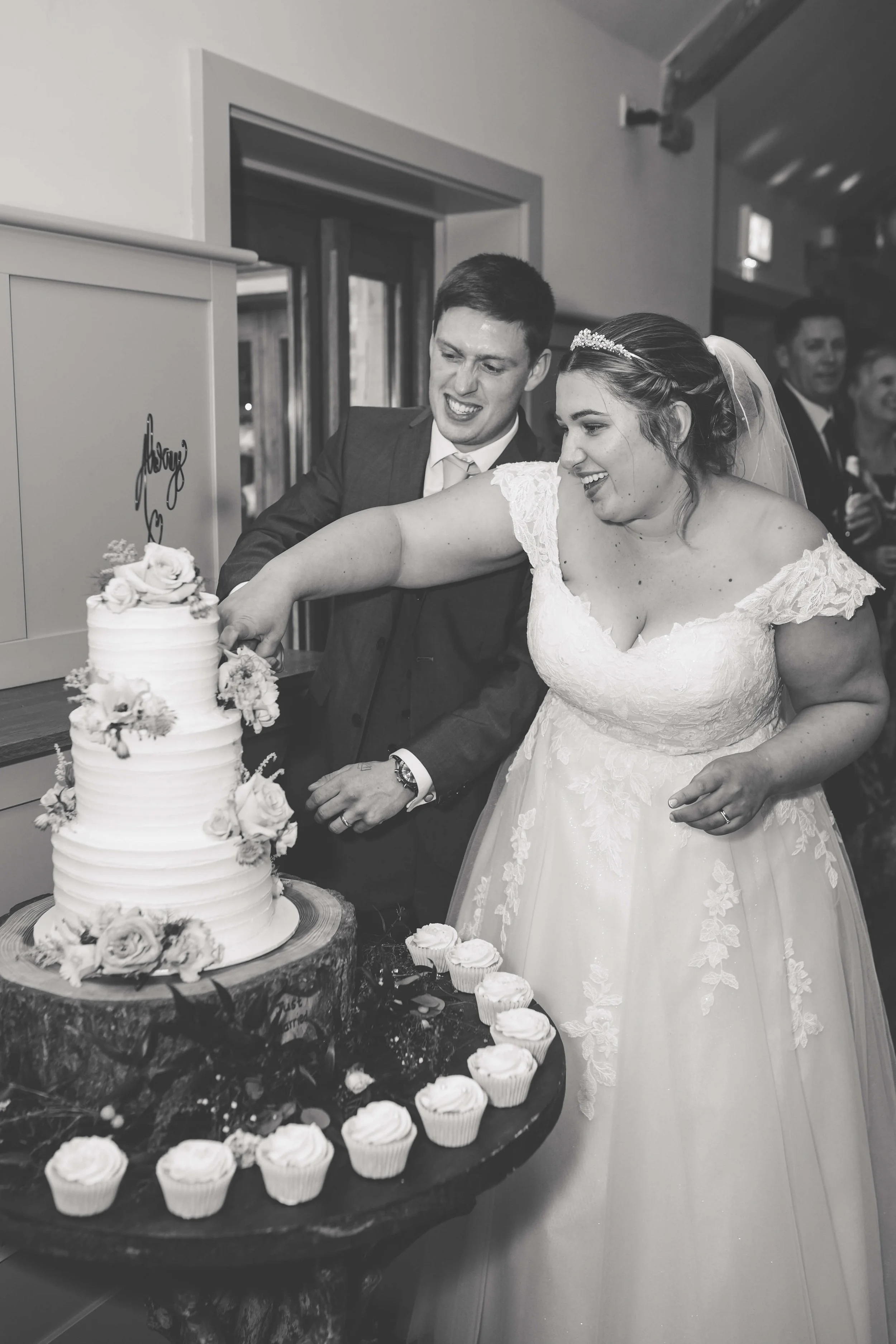 A bride and groom cut a wedding cake together at their wedding reception with guests smiling in the background.