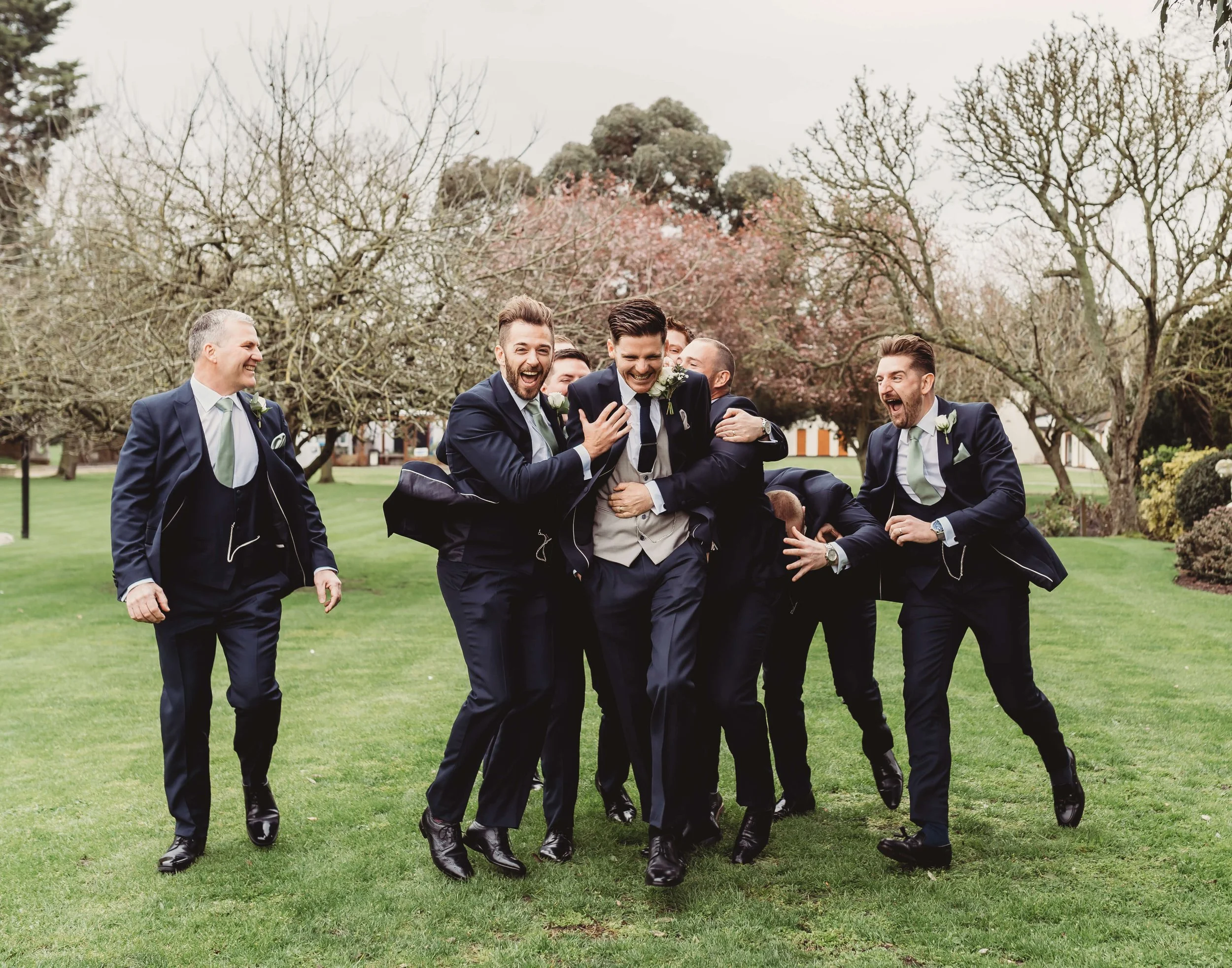 A group of groomsmen in suits celebrating and joking around outdoors on a lawn with trees in the background.