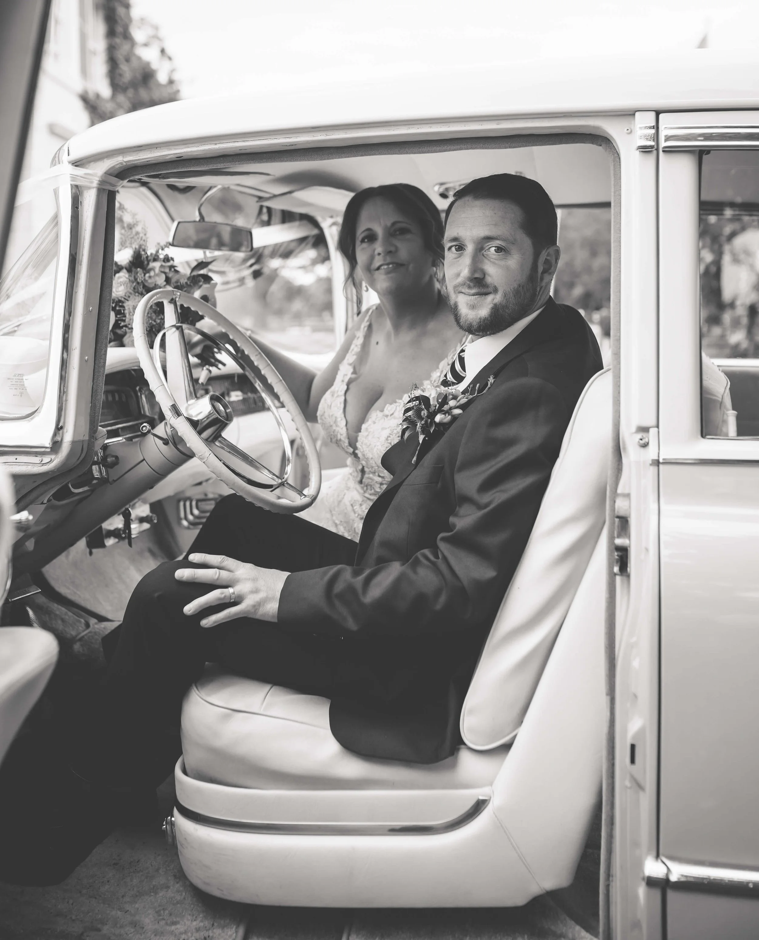 A bride and groom sitting inside a vintage car, both looking at the camera. The bride is wearing a lace wedding dress, and the groom is in a suit with a boutonniere.