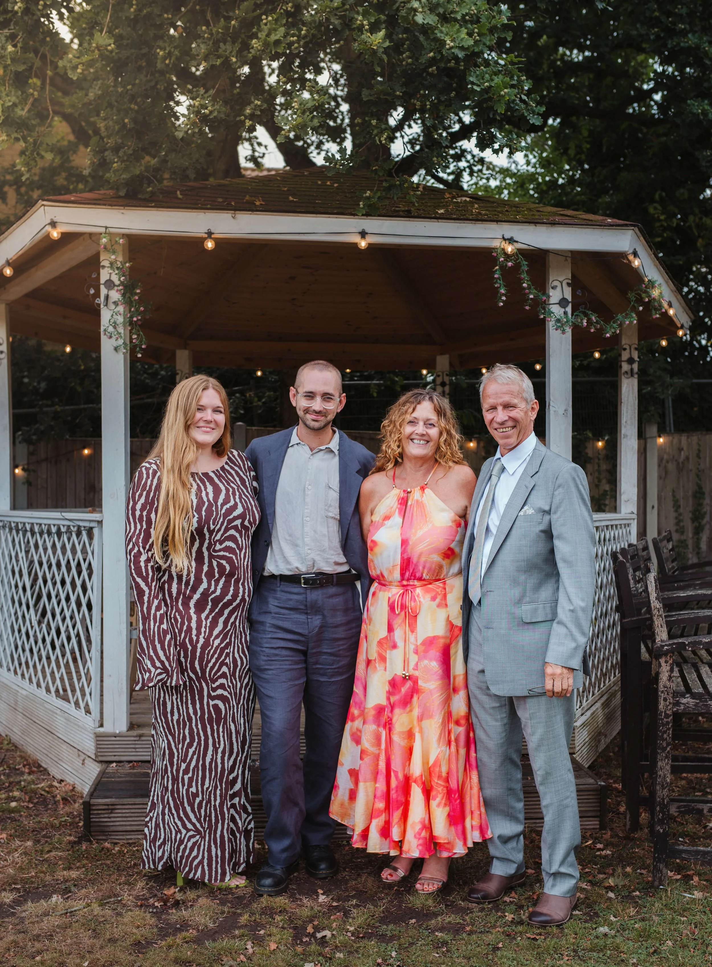 Four people standing close together in front of a gazebo, smiling, at an outdoor gathering during the evening. The group includes two women and two men dressed in casual and semi-formal attire.