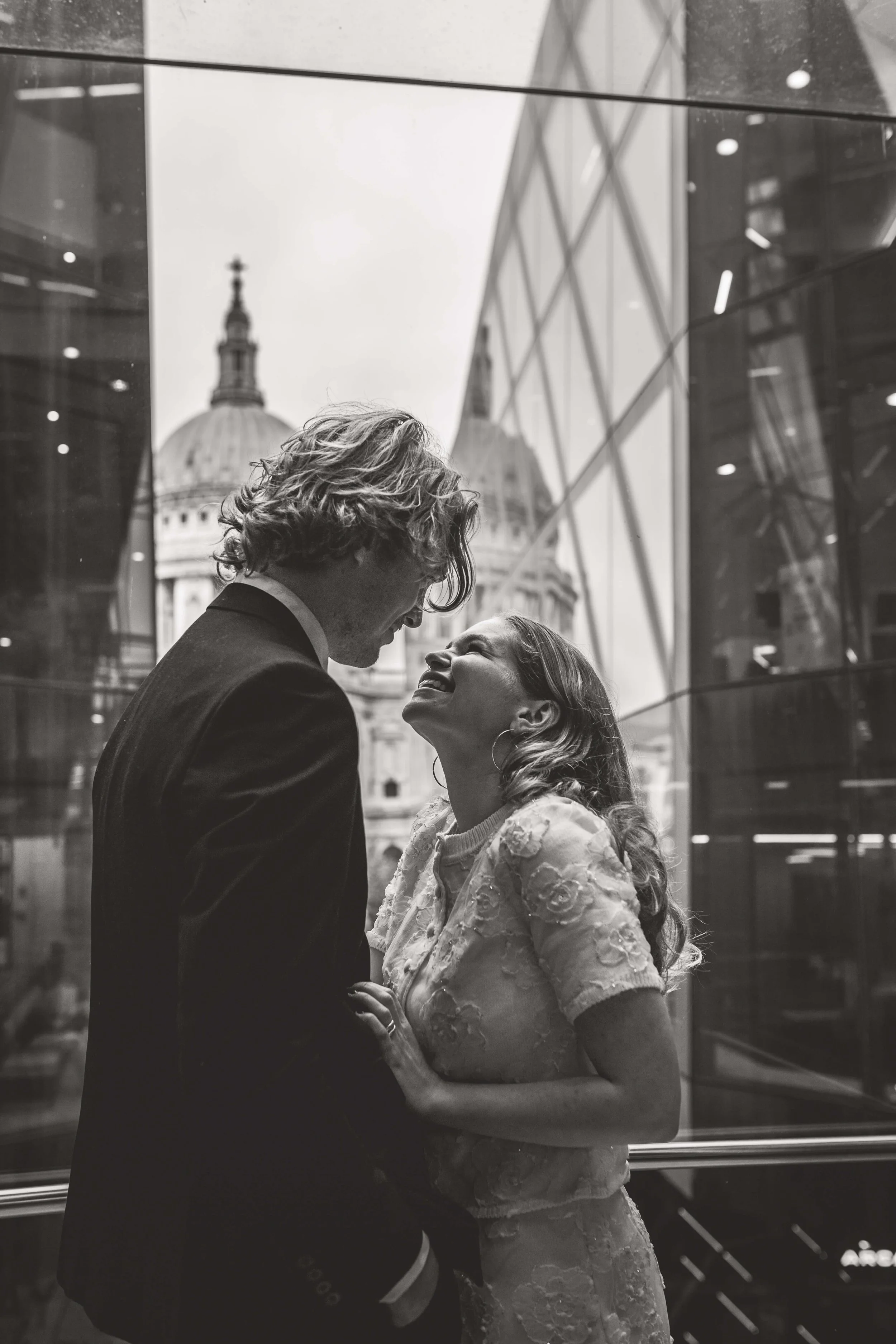 A couple dressed in wedding attire sharing a joyful moment indoors with a cityscape and classical building in the background.