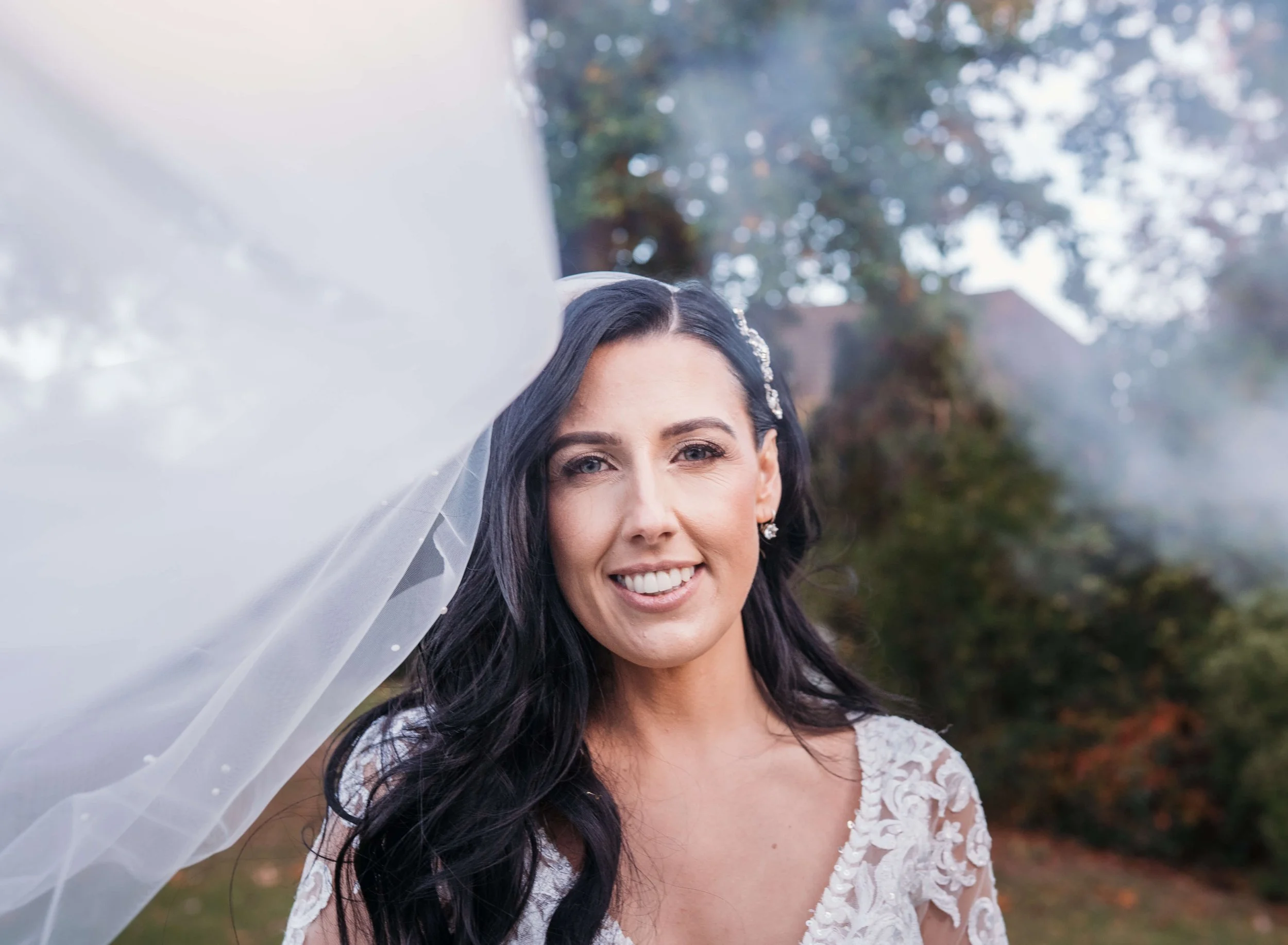 Close-up of a smiling woman with dark hair, wearing a lace wedding dress and veil, outdoors with trees and a house in the background.