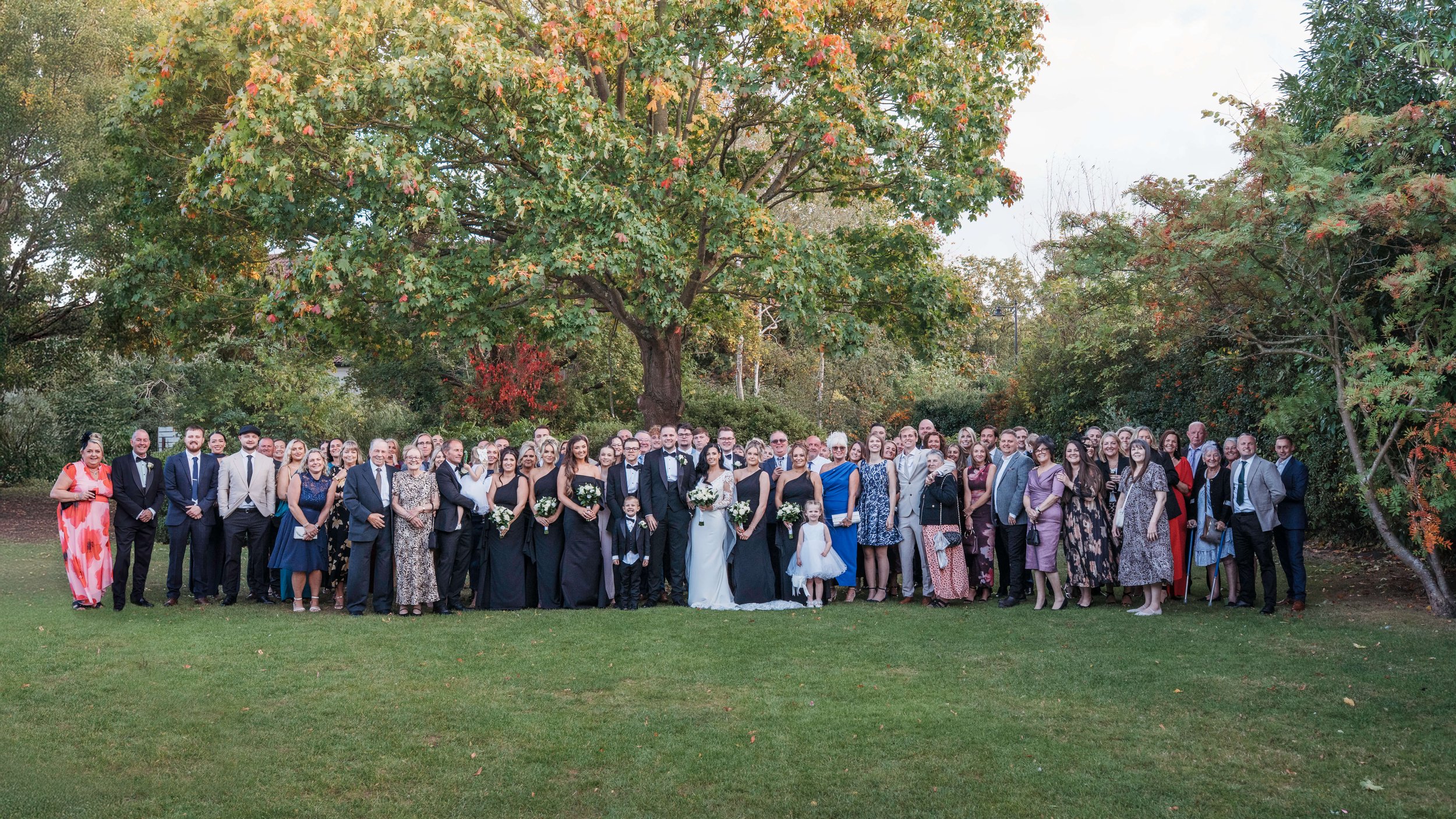 A large group of people, including the bride and groom, standing outdoors on a lawn with trees in the background, celebrating a wedding.