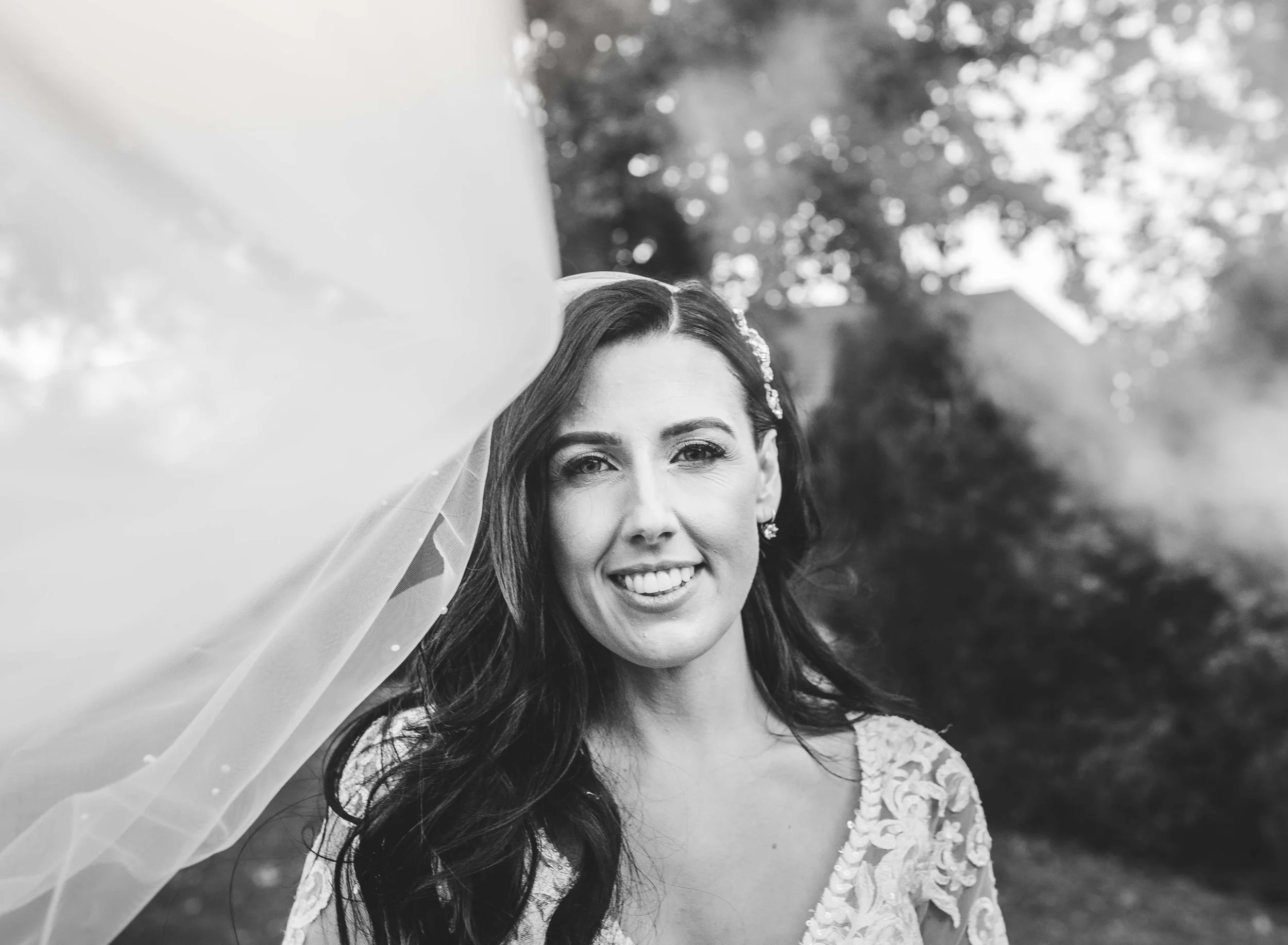 Black and white photo of a smiling woman with long dark hair, wearing a lace dress and earrings, outdoors with trees in the background.
