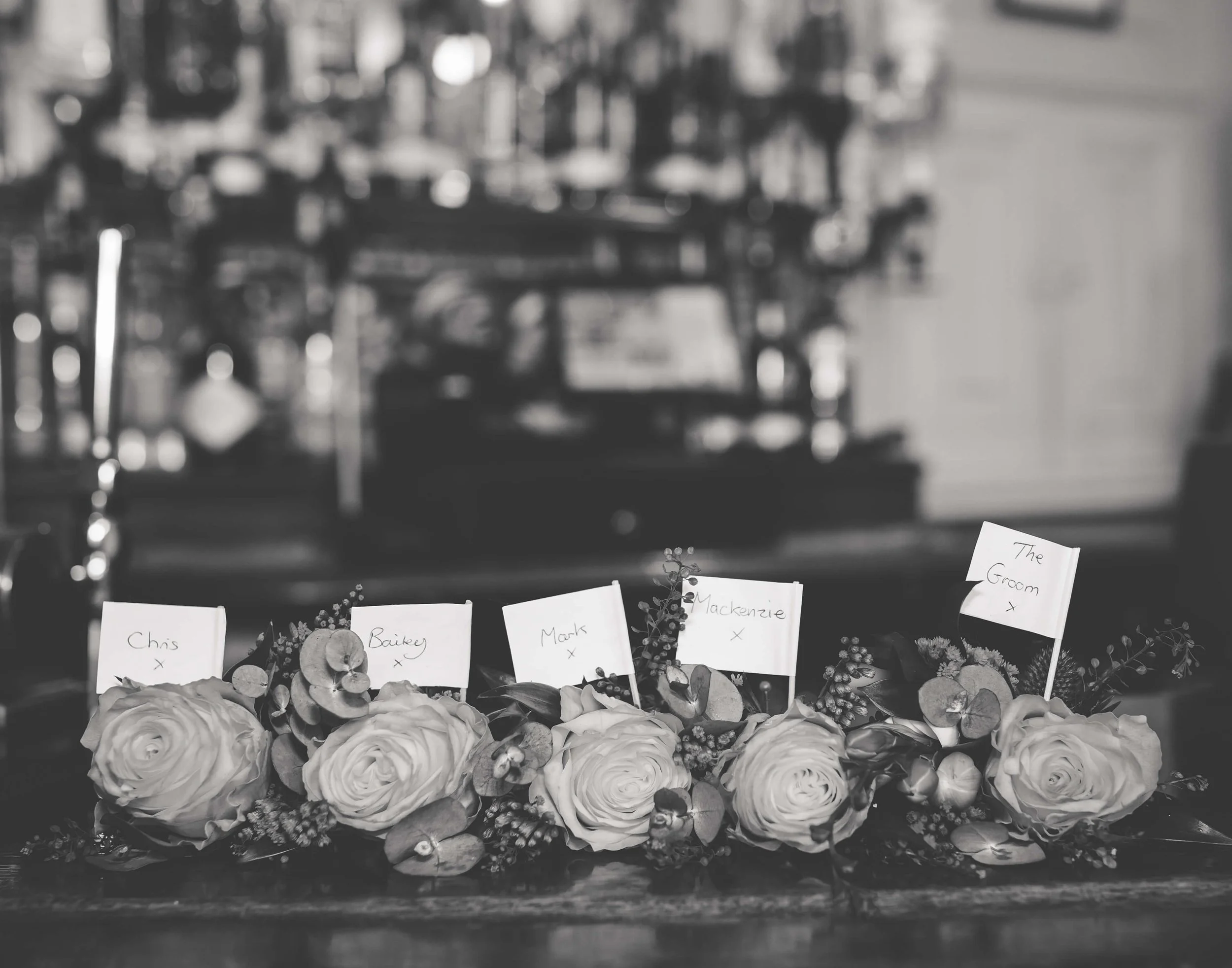 A floral arrangement with roses and greenery, featuring small flags with handwritten names including Chris, Bailey, Mark, Mackenzie, and The Groom, in a black-and-white setting.