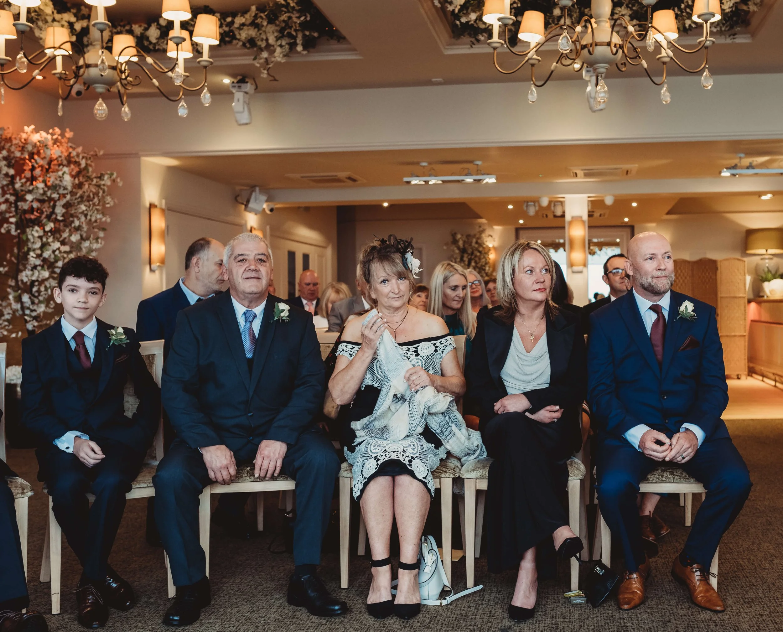 Group of people seated at a formal event, possibly a wedding, in a decorated venue with chandeliers and floral arrangements.