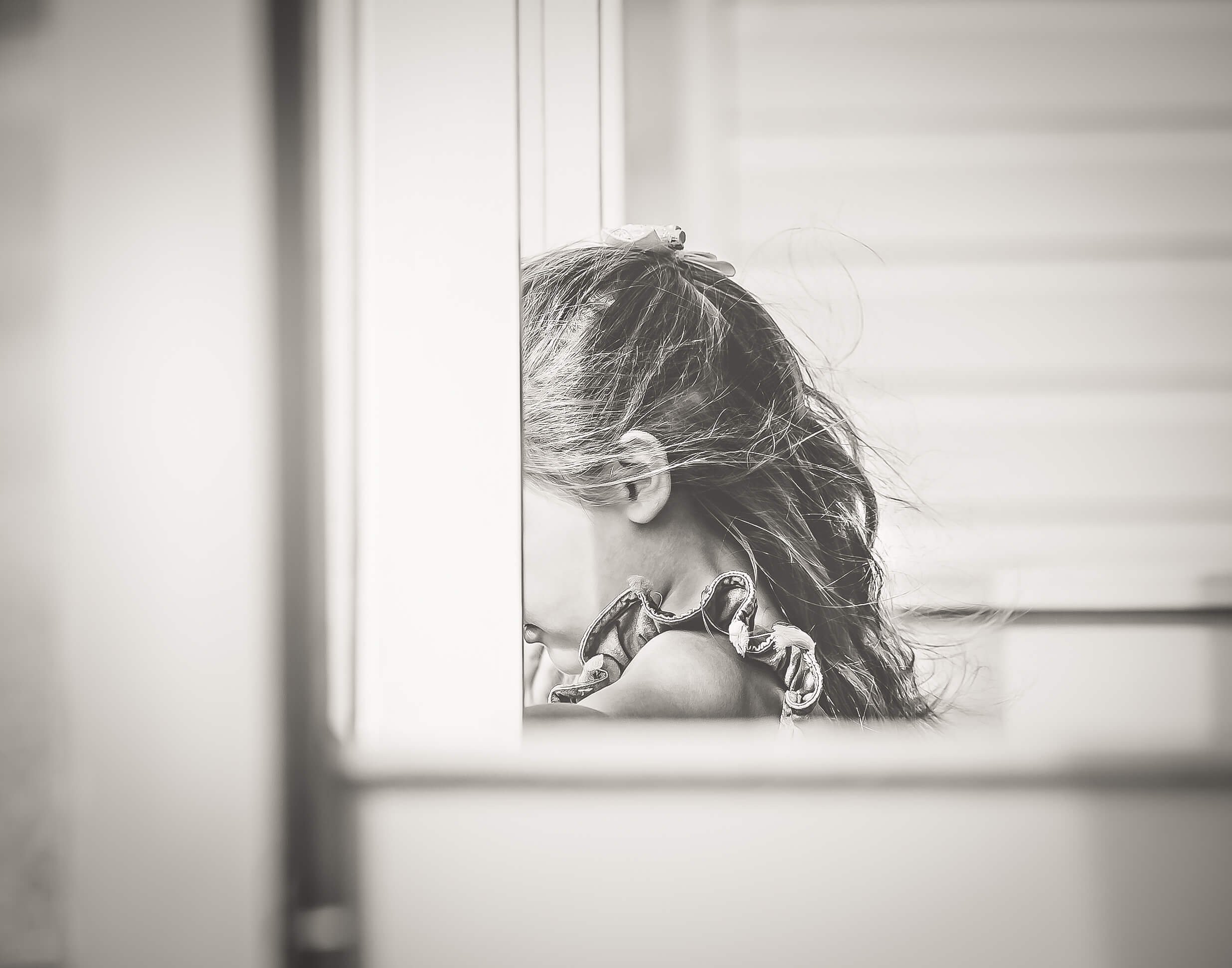 A black-and-white photo of a girl with disheveled hair, seen in profile, with her head resting on her arms. She is positioned near a window or sliding door with horizontal siding visible in the background.