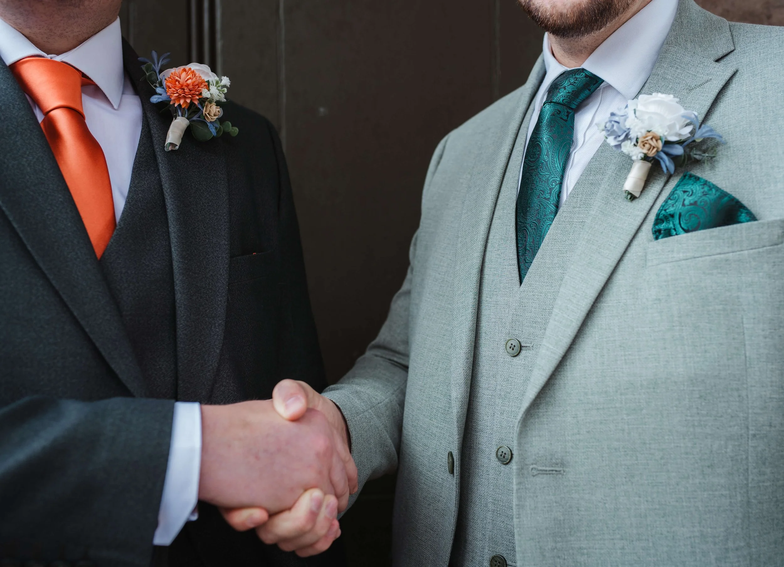 Close-up of a handshake between two men wearing suits and floral boutonnières, likely at a wedding.