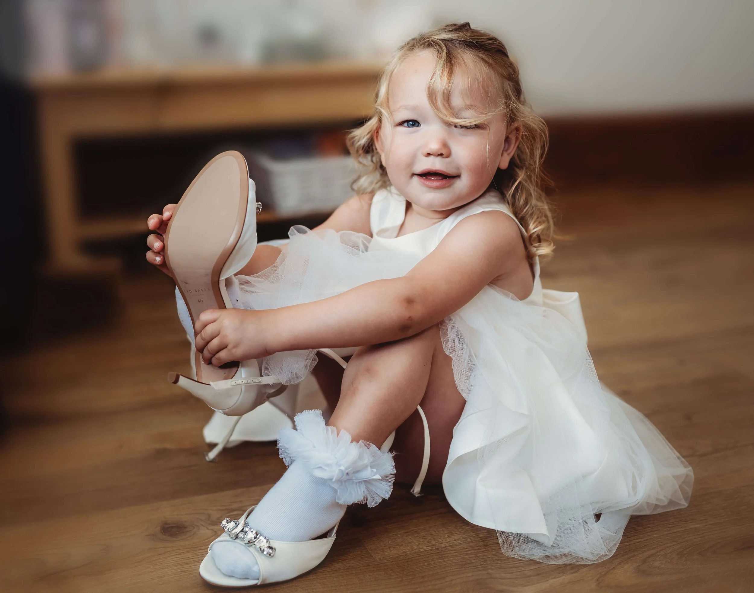 A young girl with curly blonde hair wearing a white dress and white shoes is sitting on a wooden floor, putting on a high heel shoe.