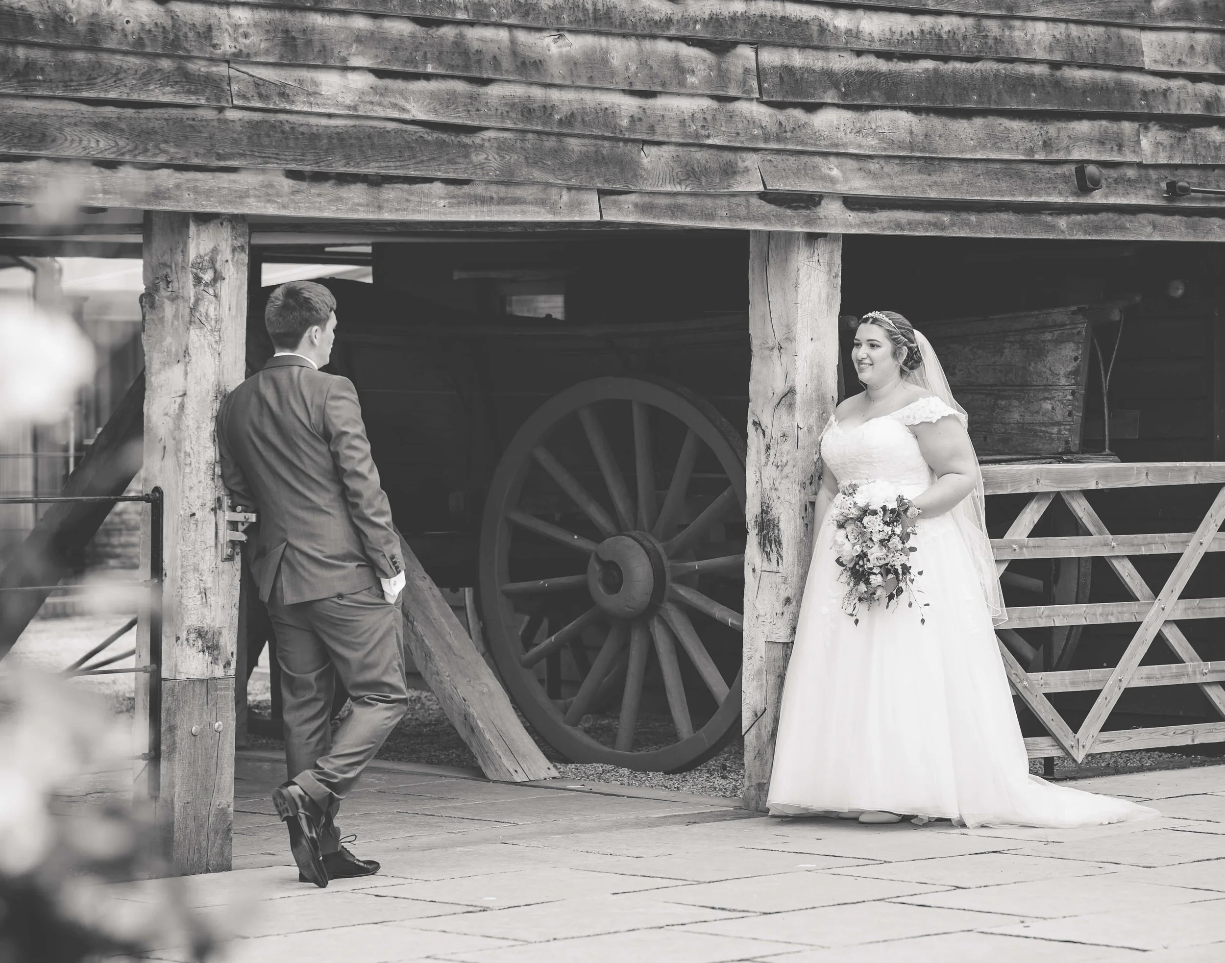 A bride and groom are standing in front of an old wooden barn, with the bride smiling and holding a bouquet of flowers, and the groom facing her with his hands in his pockets, during a wedding photoshoot.