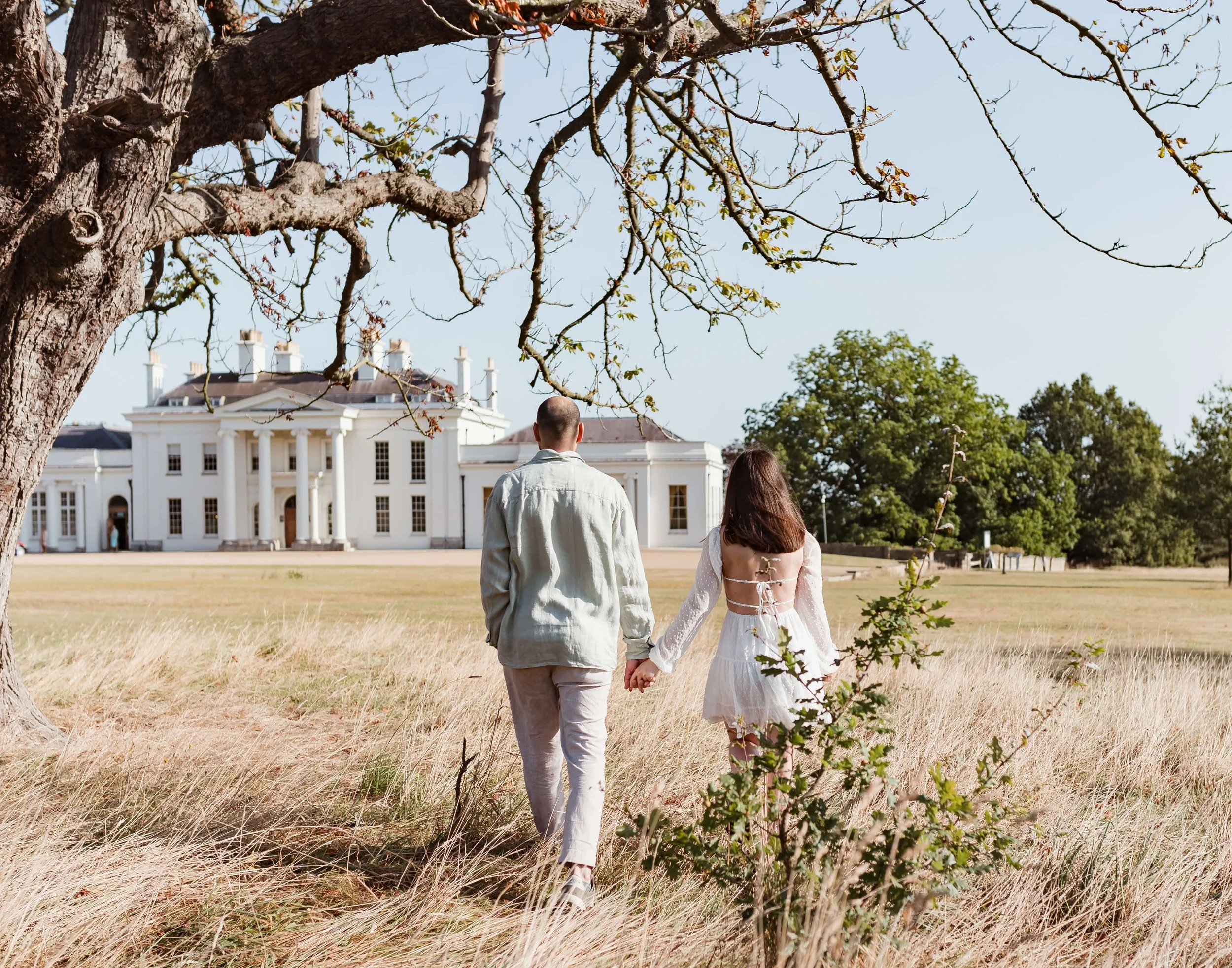 A couple holding hands walking toward a large white mansion on a sunny day, with dry grass and a large tree in the foreground.