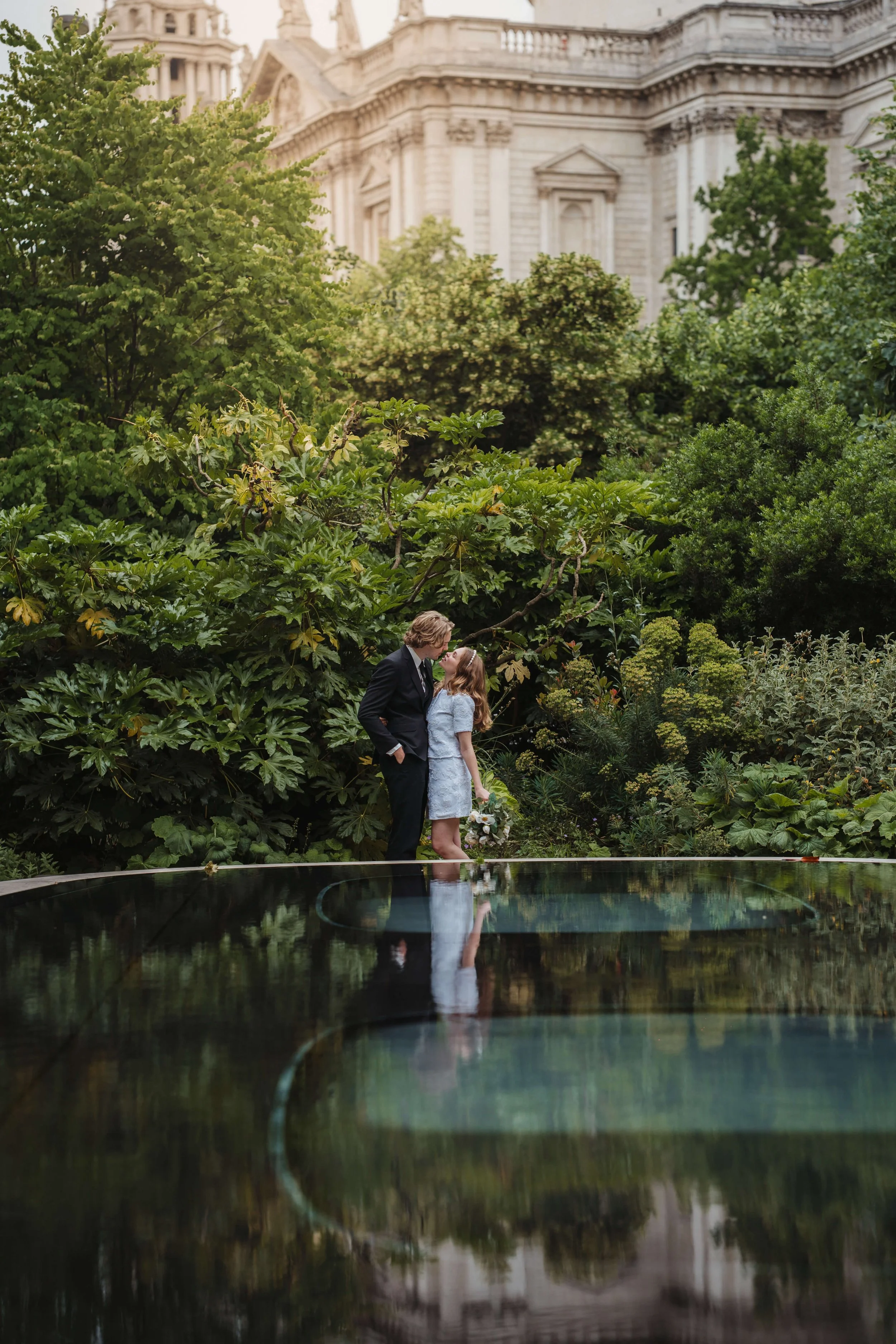 A couple sharing a kiss on a reflective surface outdoors, surrounded by lush green foliage and a historic building in the background.