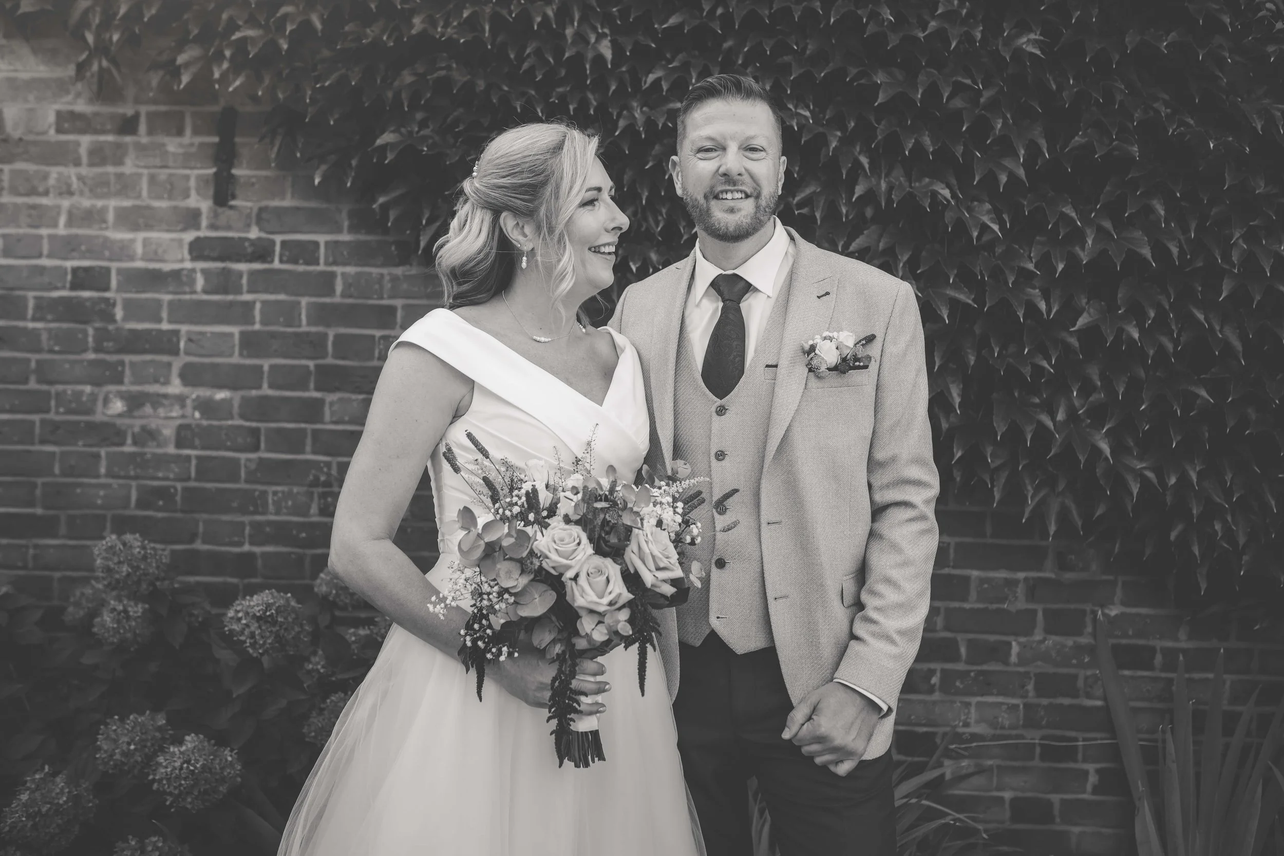 A black-and-white photo of a bride and groom standing outdoors in front of a brick wall covered in ivy. The bride, holding a bouquet of flowers, is smiling and looking at the groom. She has wavy hair and wears a sleeveless wedding dress. The groom, w