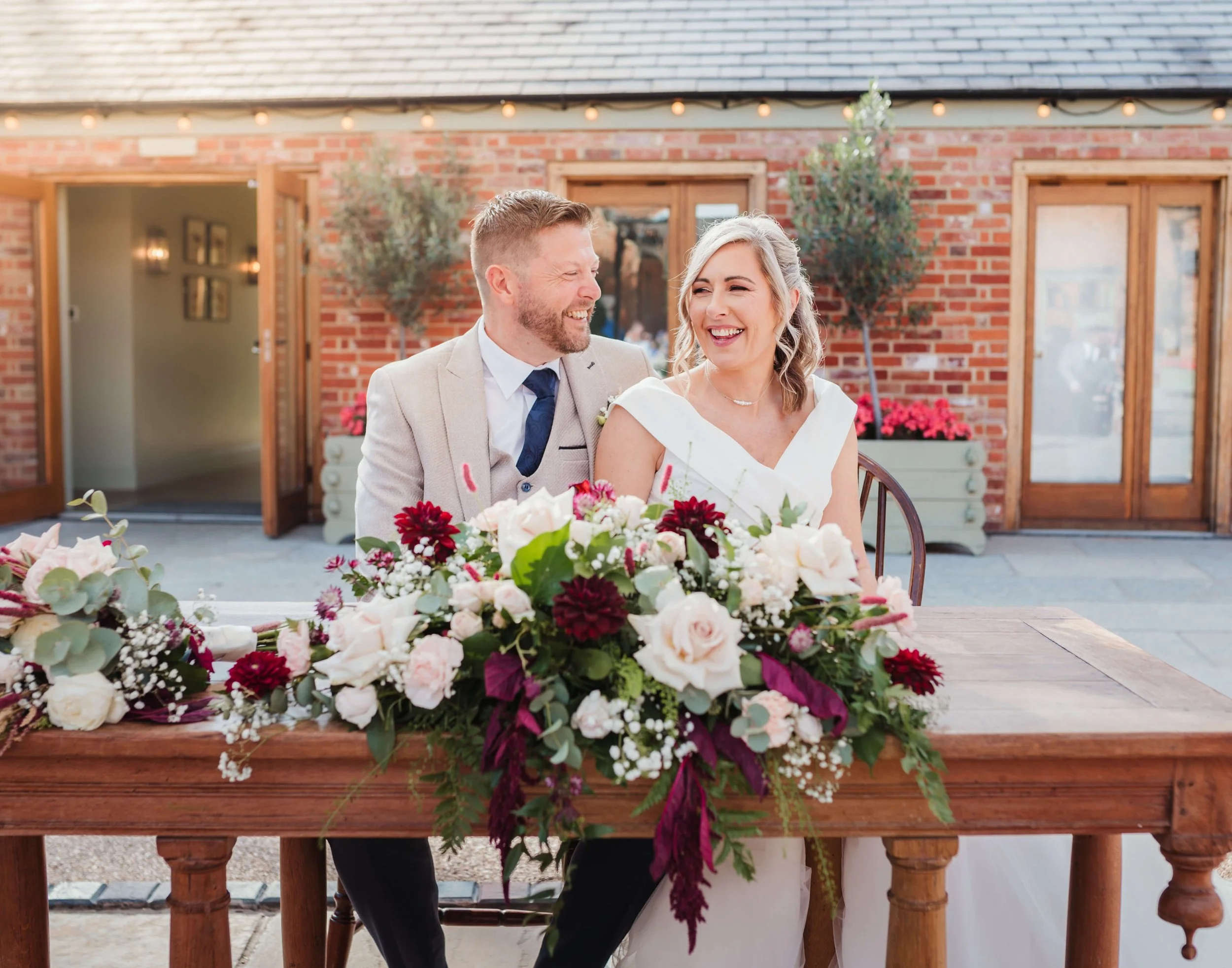 A smiling newlywed couple sitting at a table decorated with a large floral arrangement in an outdoor venue.