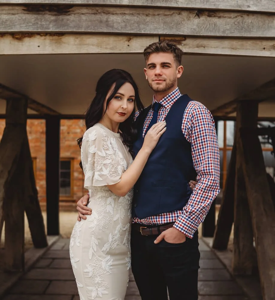 A young couple standing together under a wooden structure, with the woman in a white embroidered dress and the man in a plaid shirt with a vest, posing for a photo outdoors.